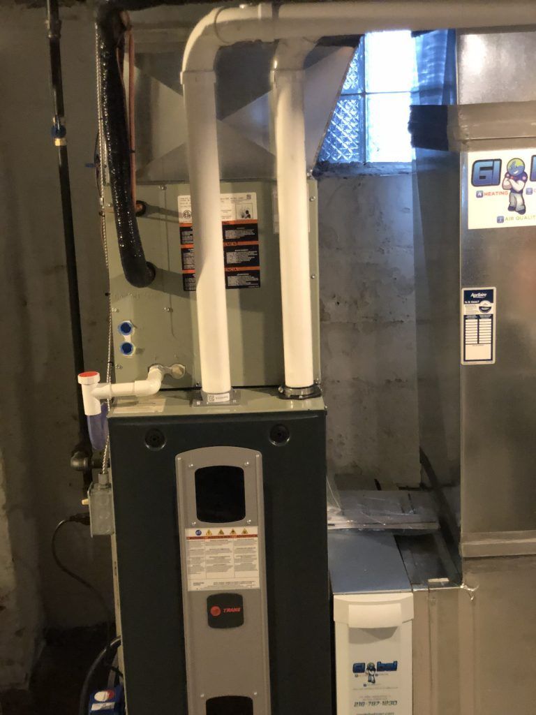 A furnace in a basement with white pipes, a gray metal exterior, and a white filter.