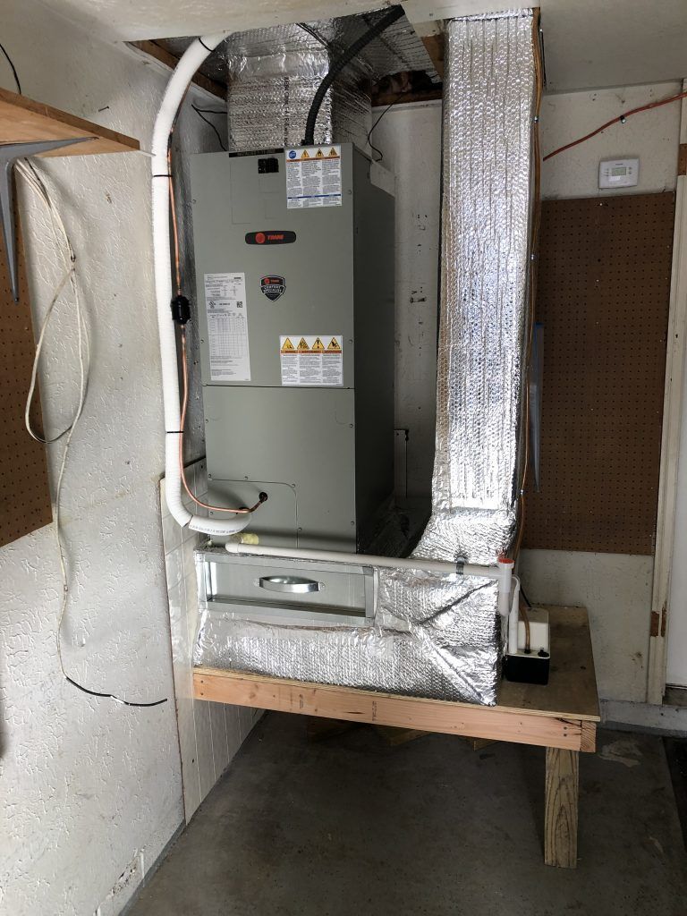 Furnace unit in a garage, with ductwork wrapped in foil insulation and a white condensation drain pipe.