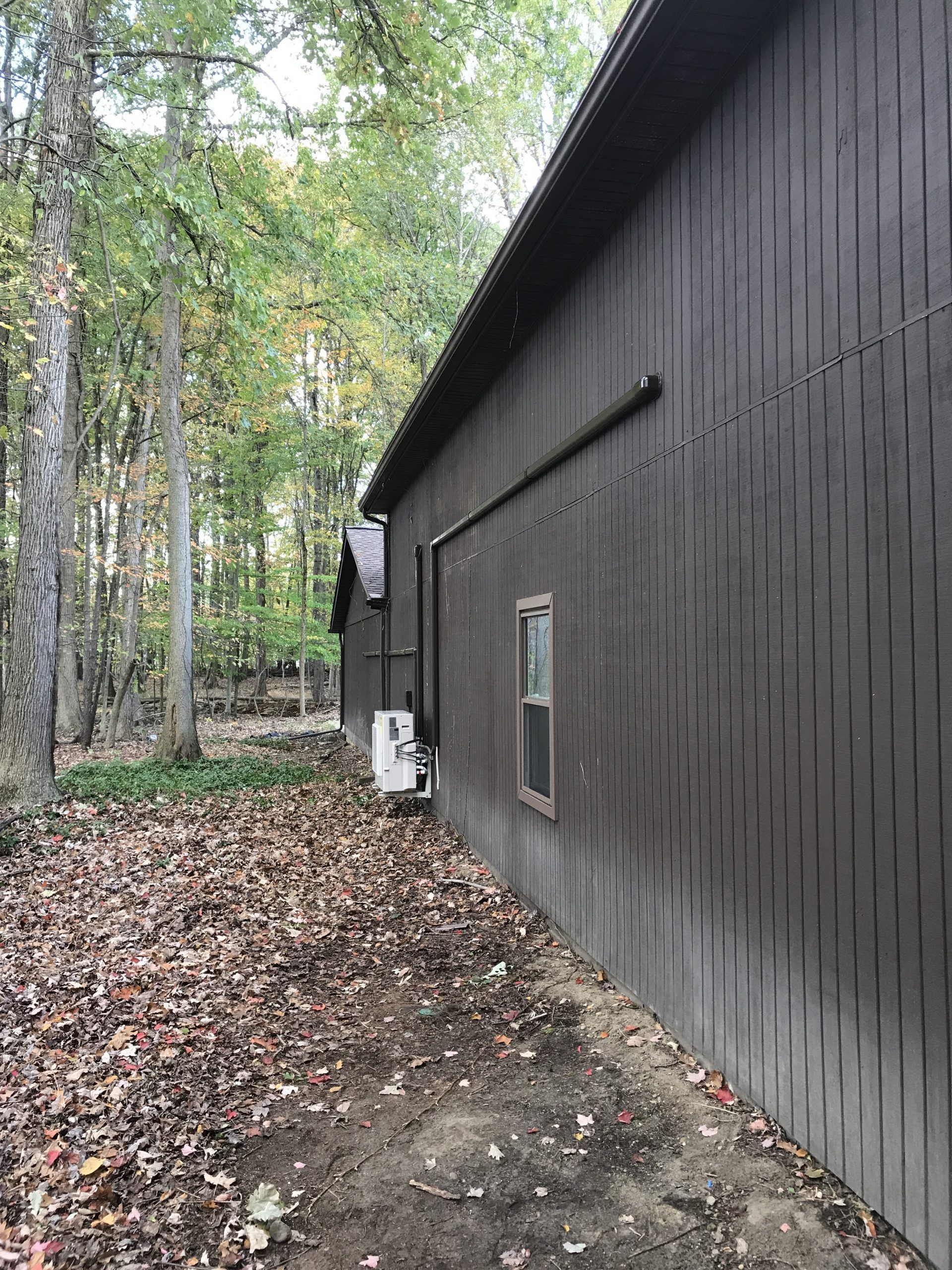 Brown building exterior with a window and a black pipe on a wooded property with fallen leaves.