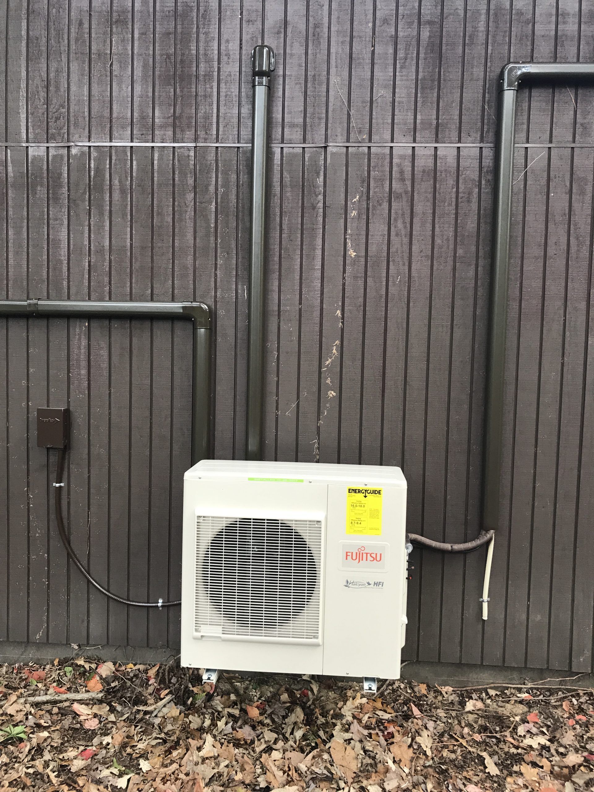 White air conditioning unit mounted on a gray wooden wall with attached pipes and electrical box.