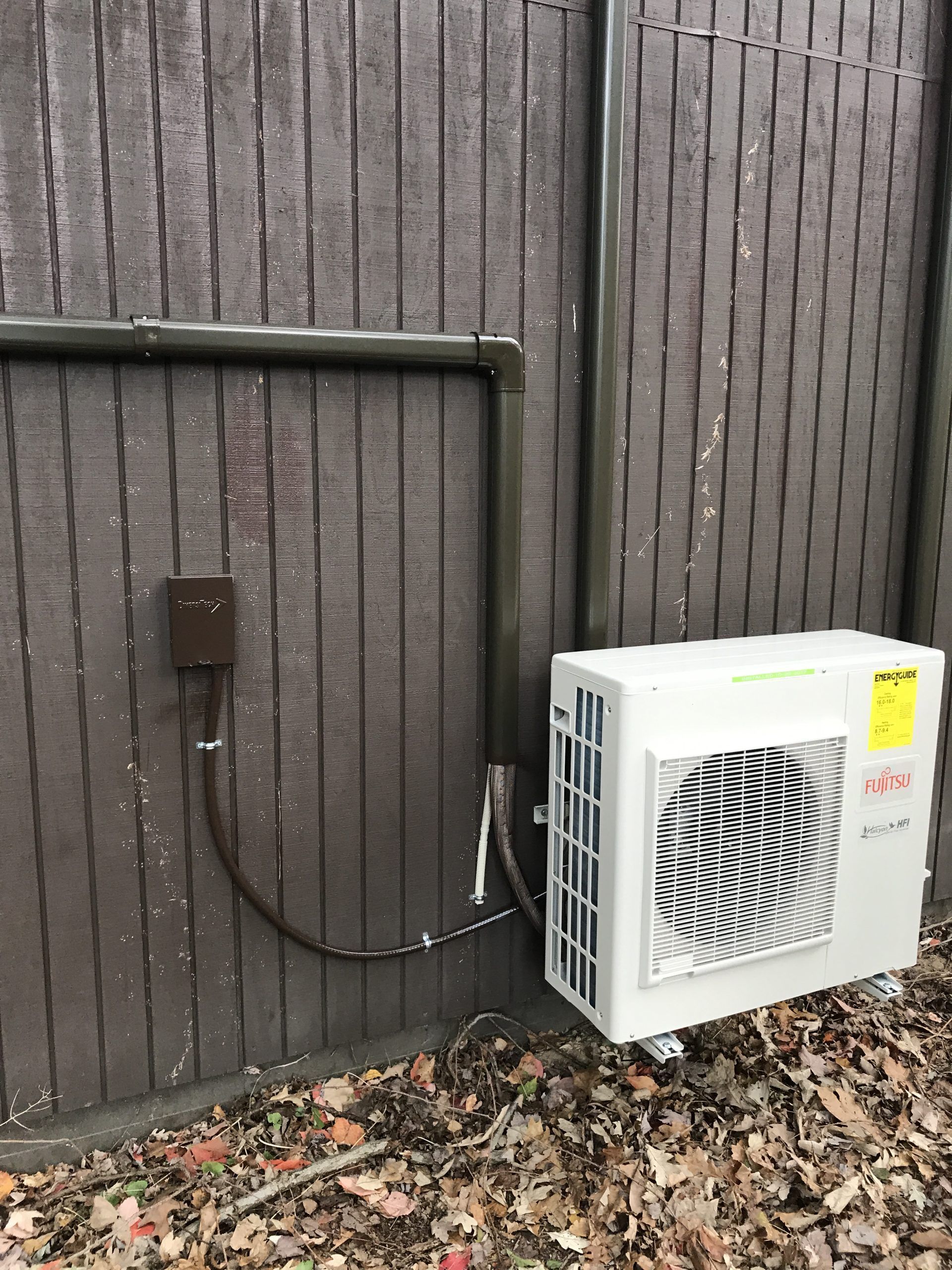 An outdoor air conditioning unit with black pipes on a brown wooden wall. A small electrical box is visible.