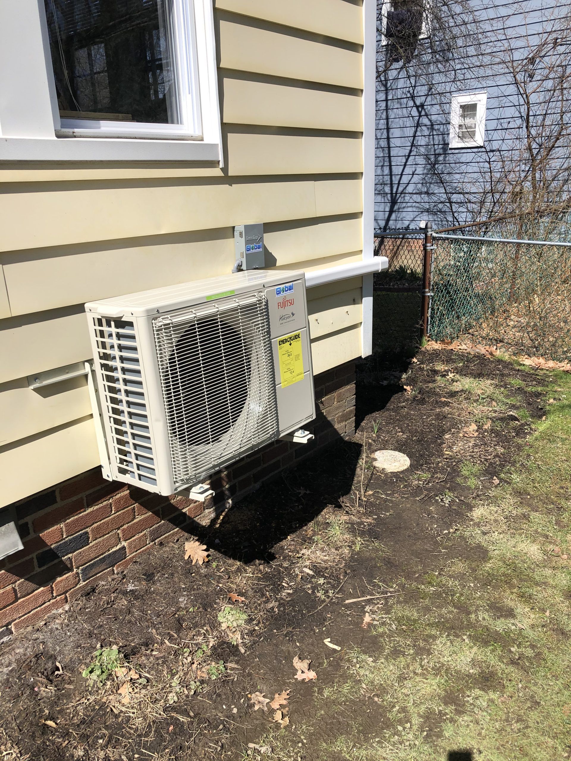 Outdoor HVAC unit mounted on a light yellow house with a window, on dirt and grass.