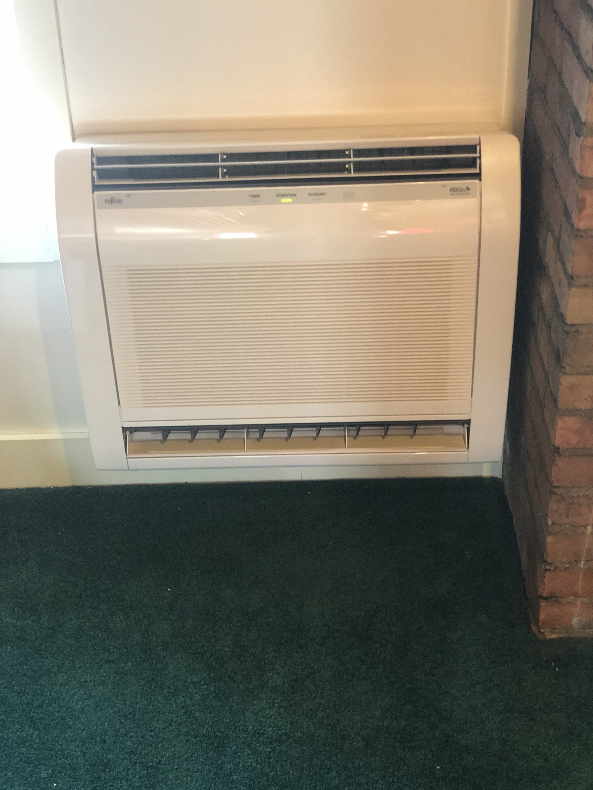 White, wall-mounted air conditioning unit against a white wall and green carpet next to a brick wall.