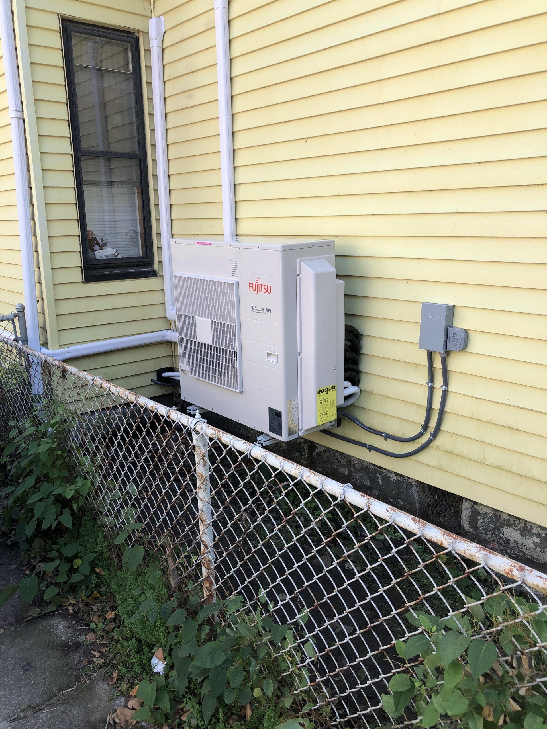 Exterior heat pump unit on a yellow house. White pipes, chain-link fence, and greenery are visible.