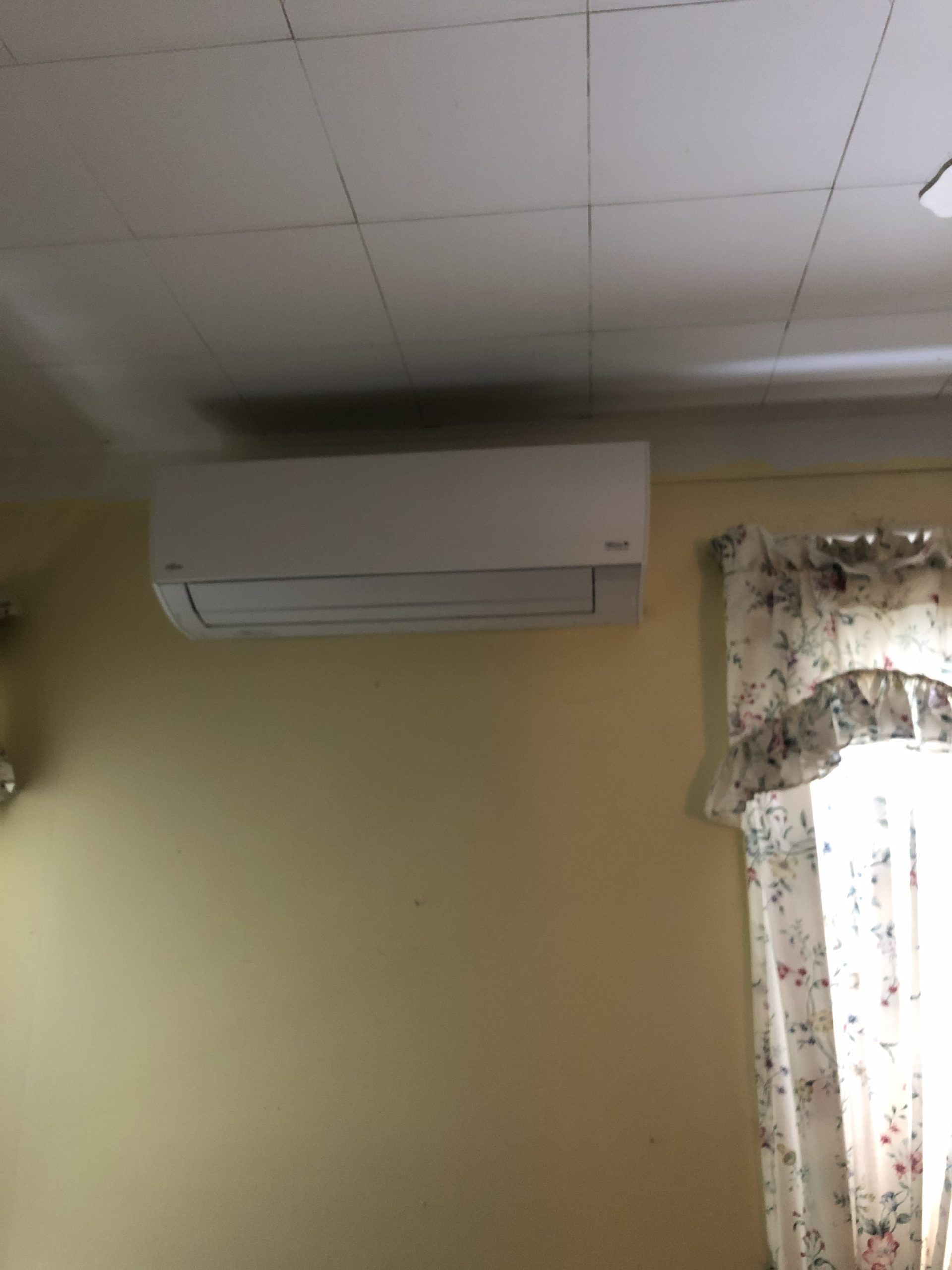 White wall-mounted air conditioner on a yellow wall, near a window with floral curtains.