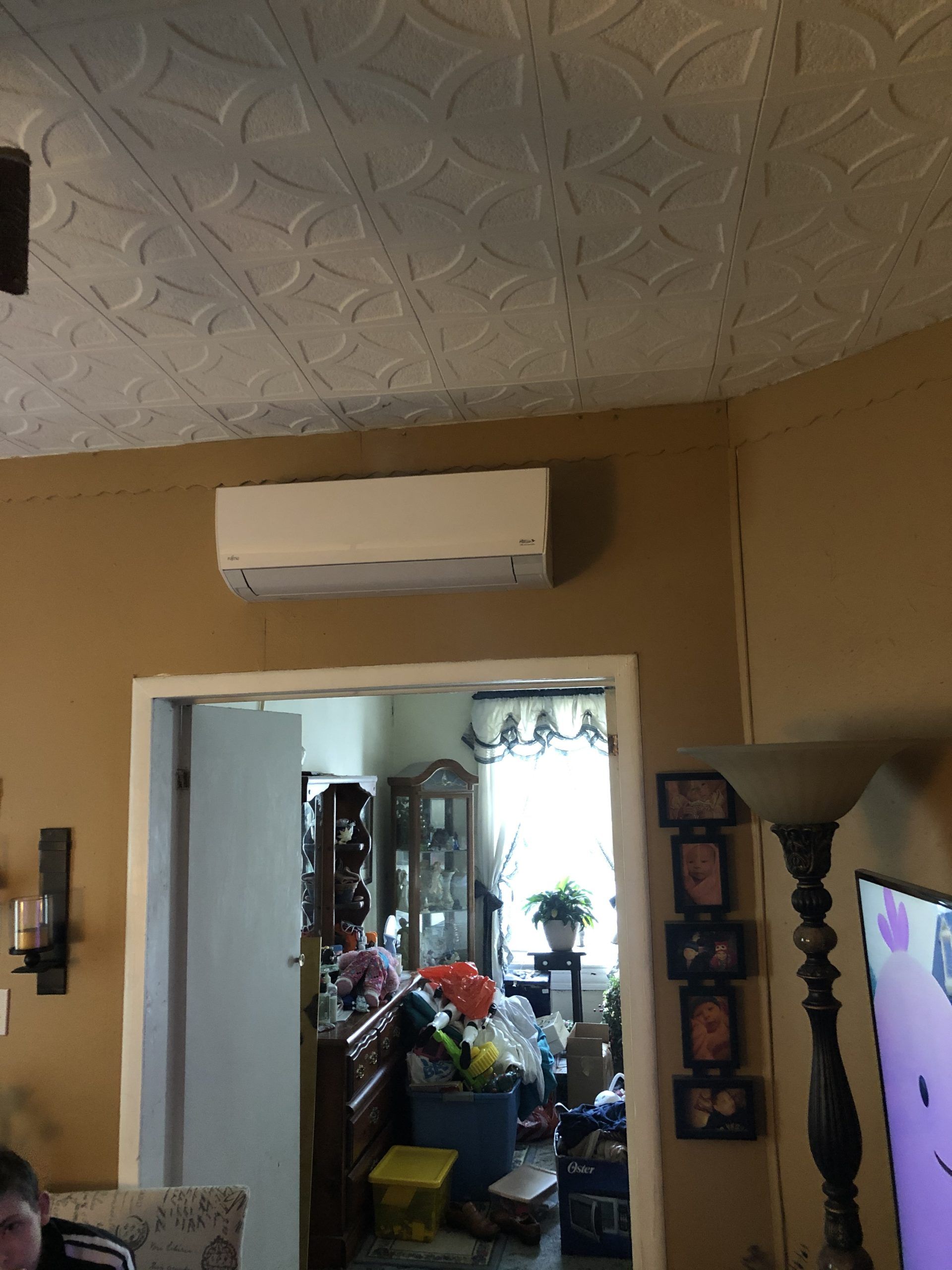 Wall-mounted air conditioning unit above a doorway, on a tan wall. The room beyond is cluttered.