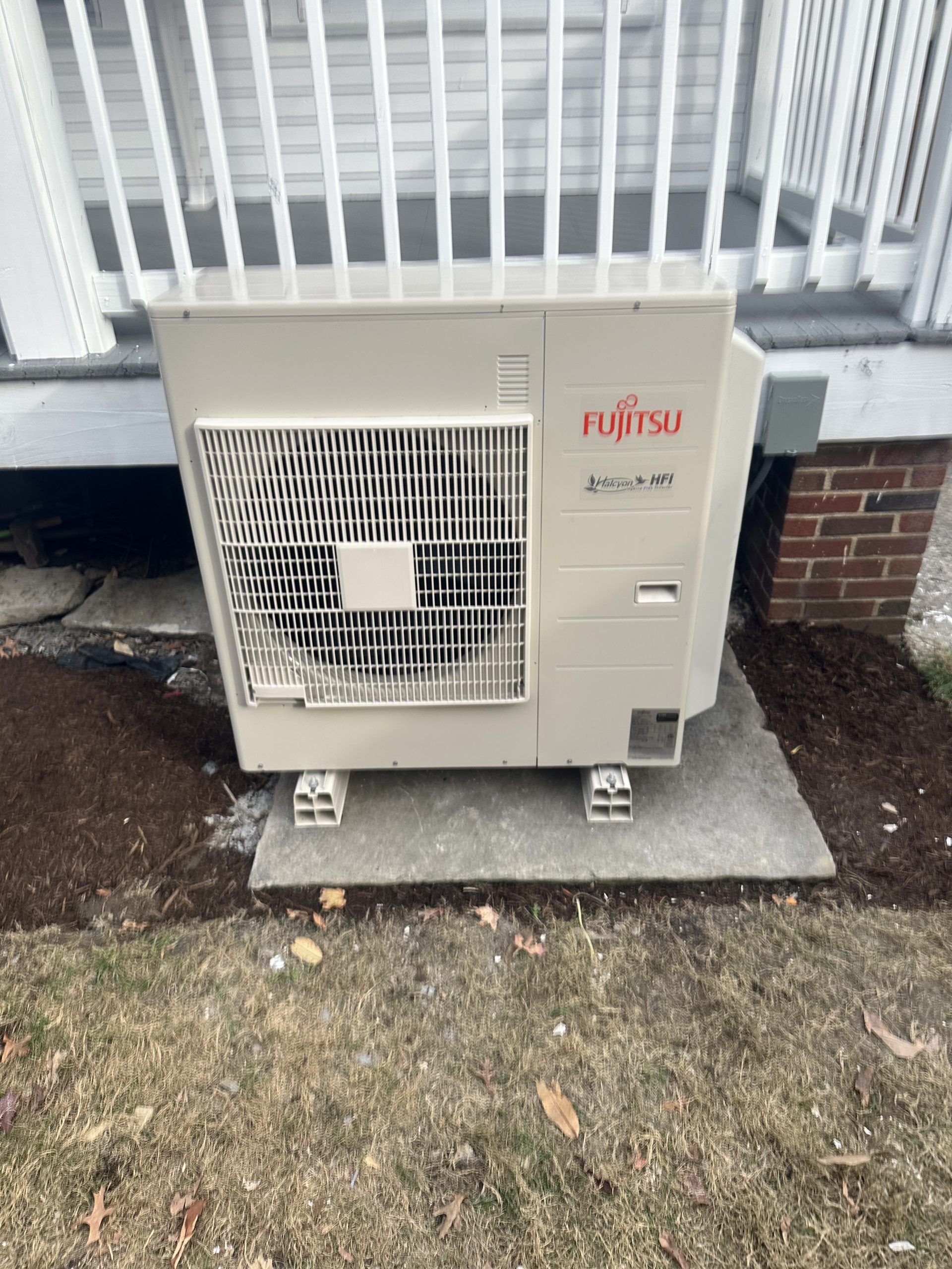 Fujitsu heat pump unit, beige, on concrete base, next to brick foundation.