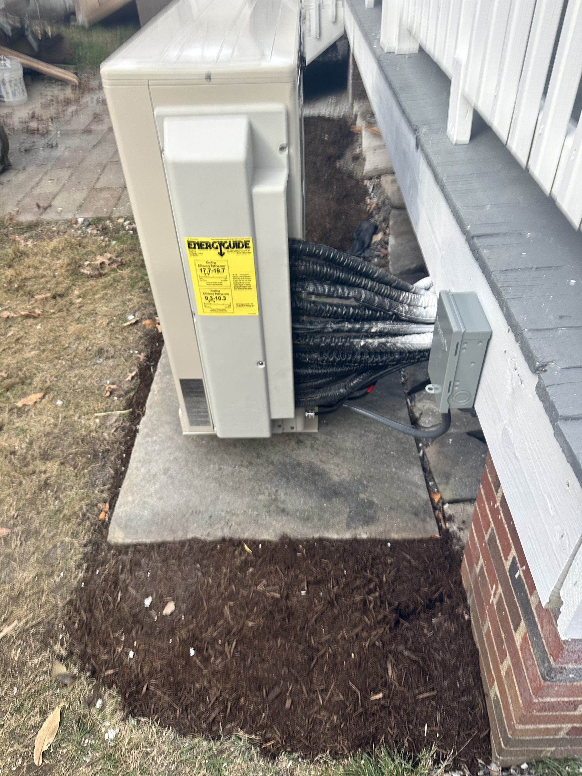 An outdoor heat pump unit on a concrete pad next to a house with black tubing extending out, and mulch around the base.