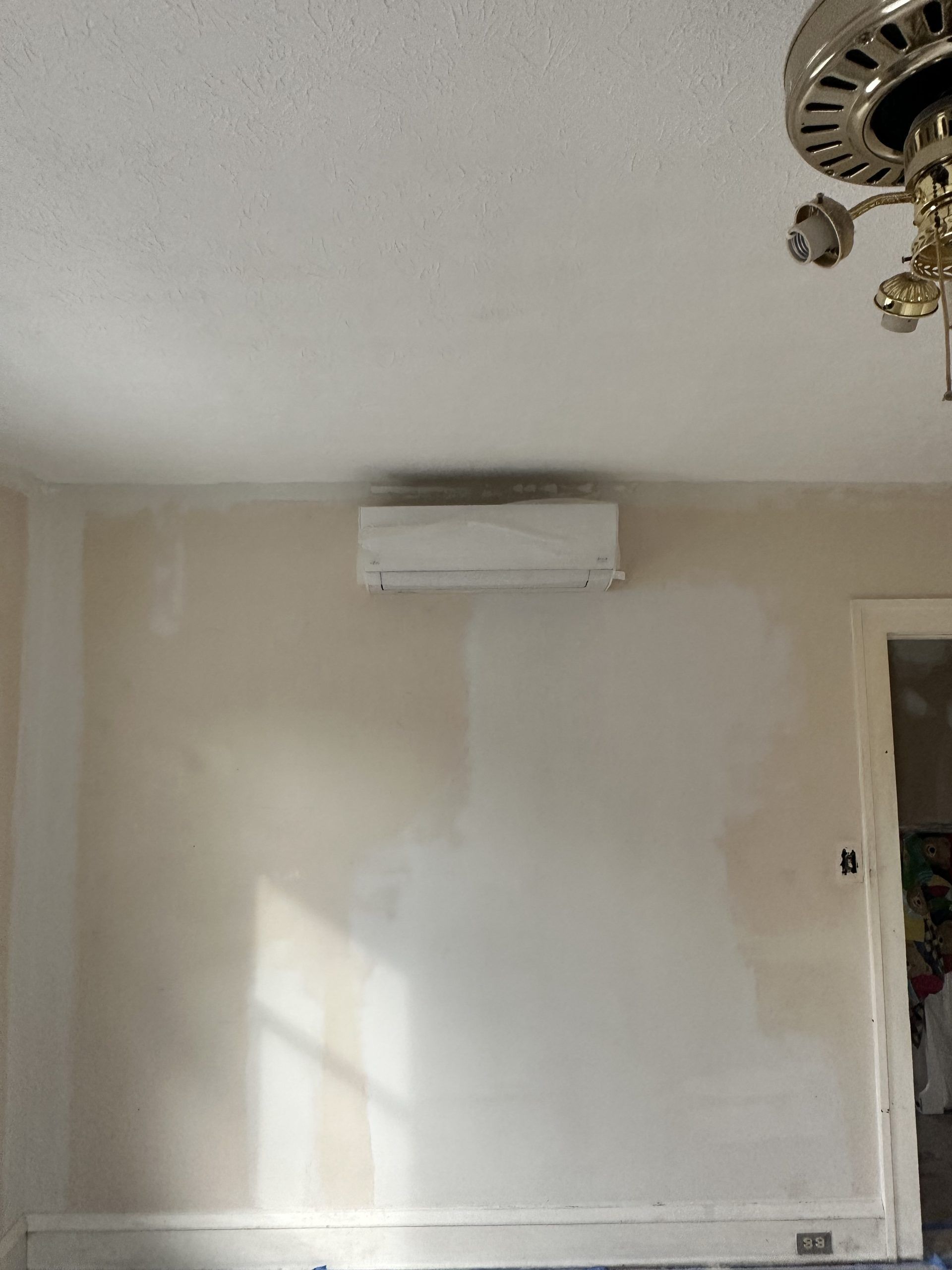White wall-mounted air conditioning unit on a beige wall in a room. Ceiling fan on the right.