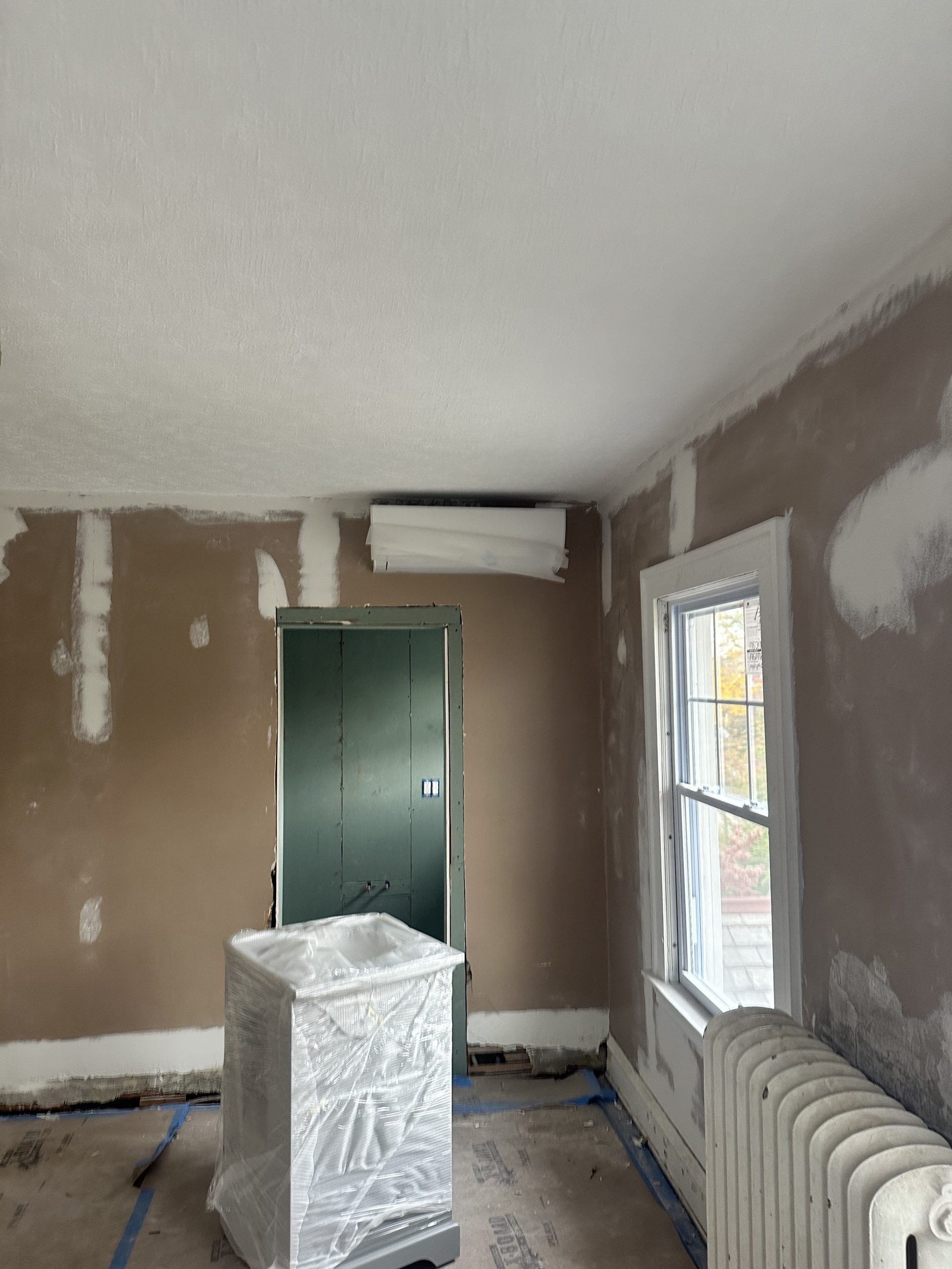 Room under renovation; walls and ceiling plastered, green door, covered appliance.