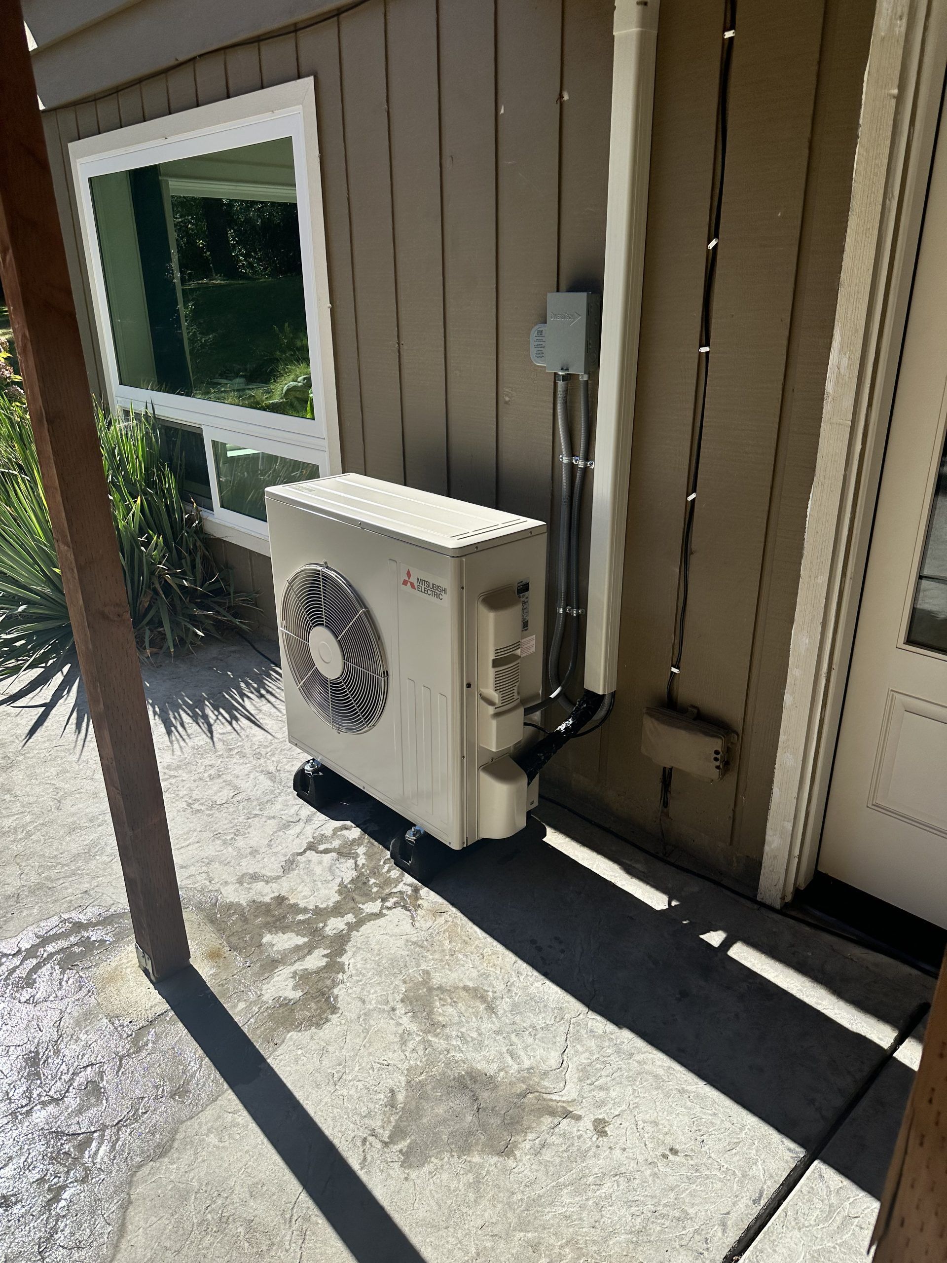 An exterior HVAC unit against a beige house with a window and a door.