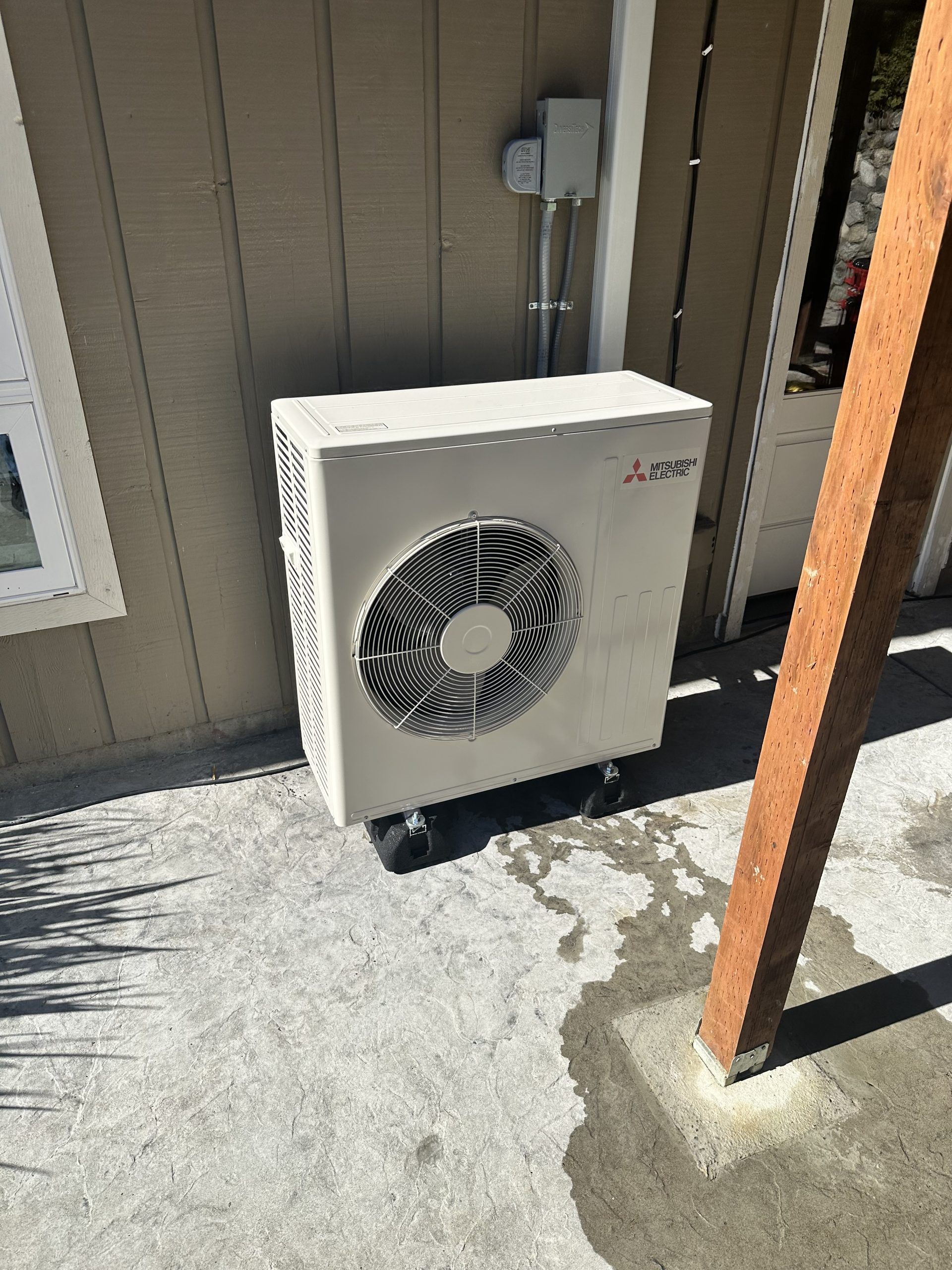 Mitsubishi heat pump unit, beige, mounted on a concrete patio next to a house wall and wooden post.