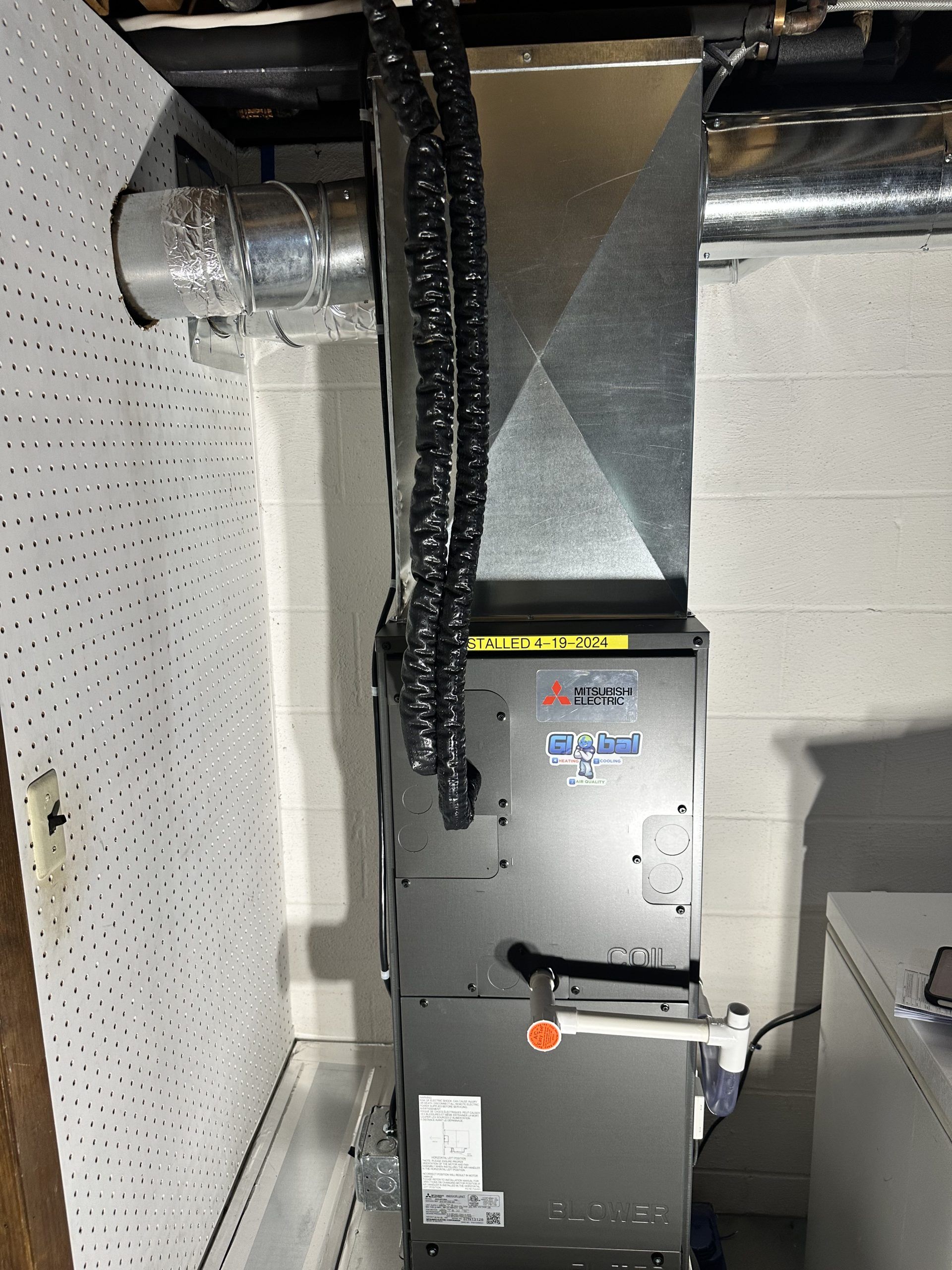 Furnace unit in a utility room with connected ductwork and pipes against a white cinder block wall.