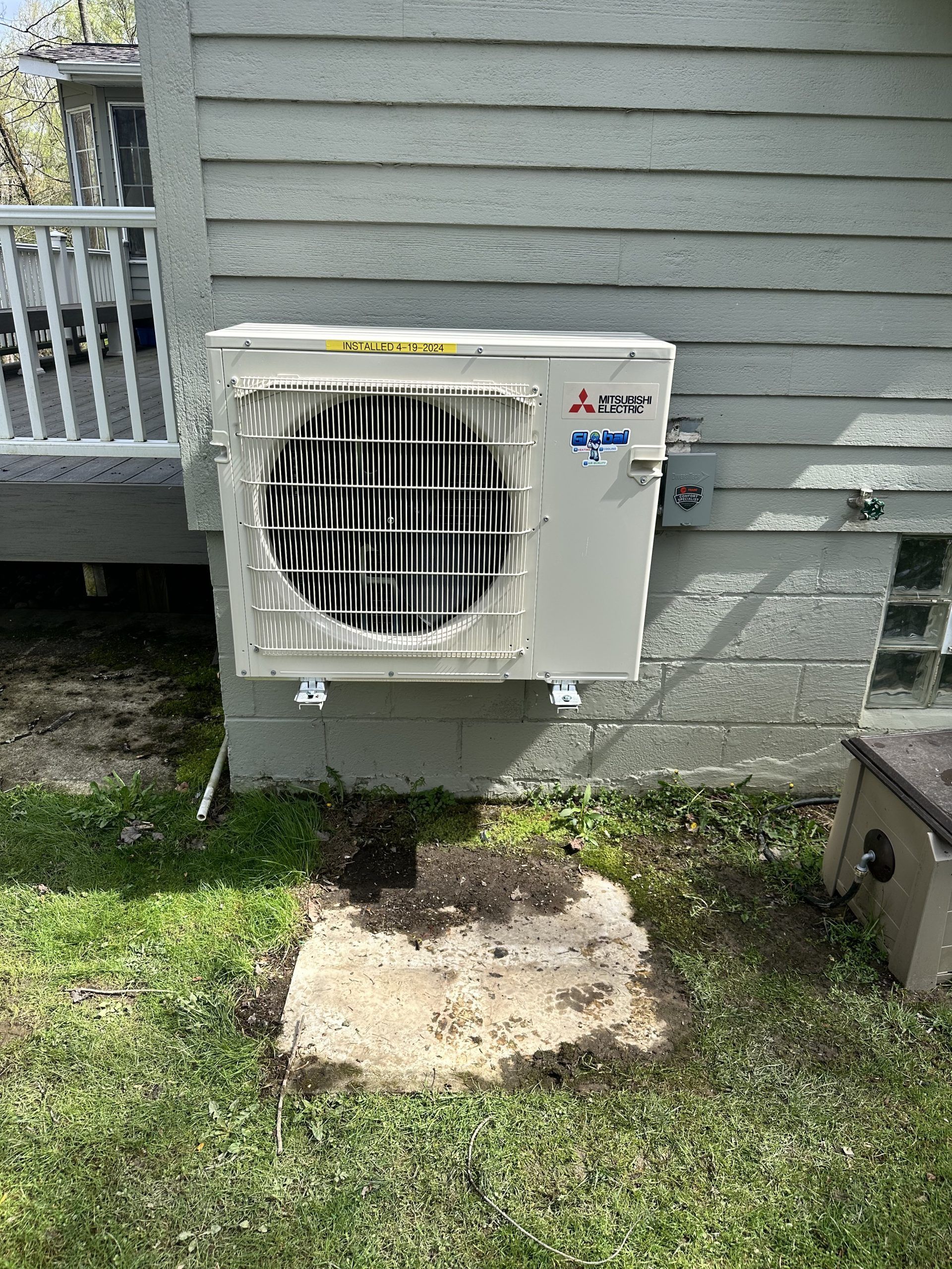 Outdoor air conditioning unit mounted on a light gray house. Grassy area with a concrete square in the foreground.