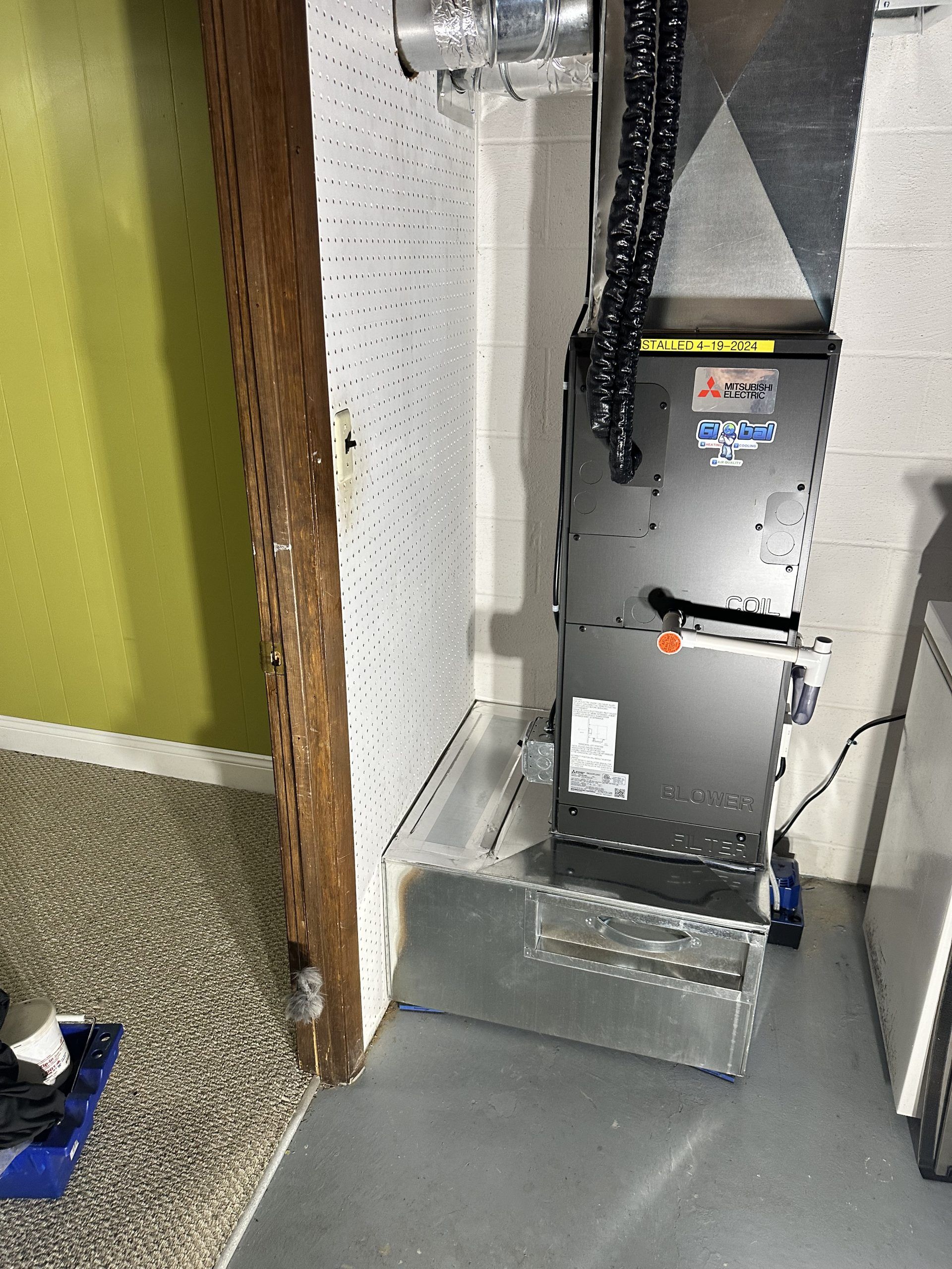 HVAC system in a basement.  A silver metal furnace with ducts, pipes, and electrical cords against a light wall.