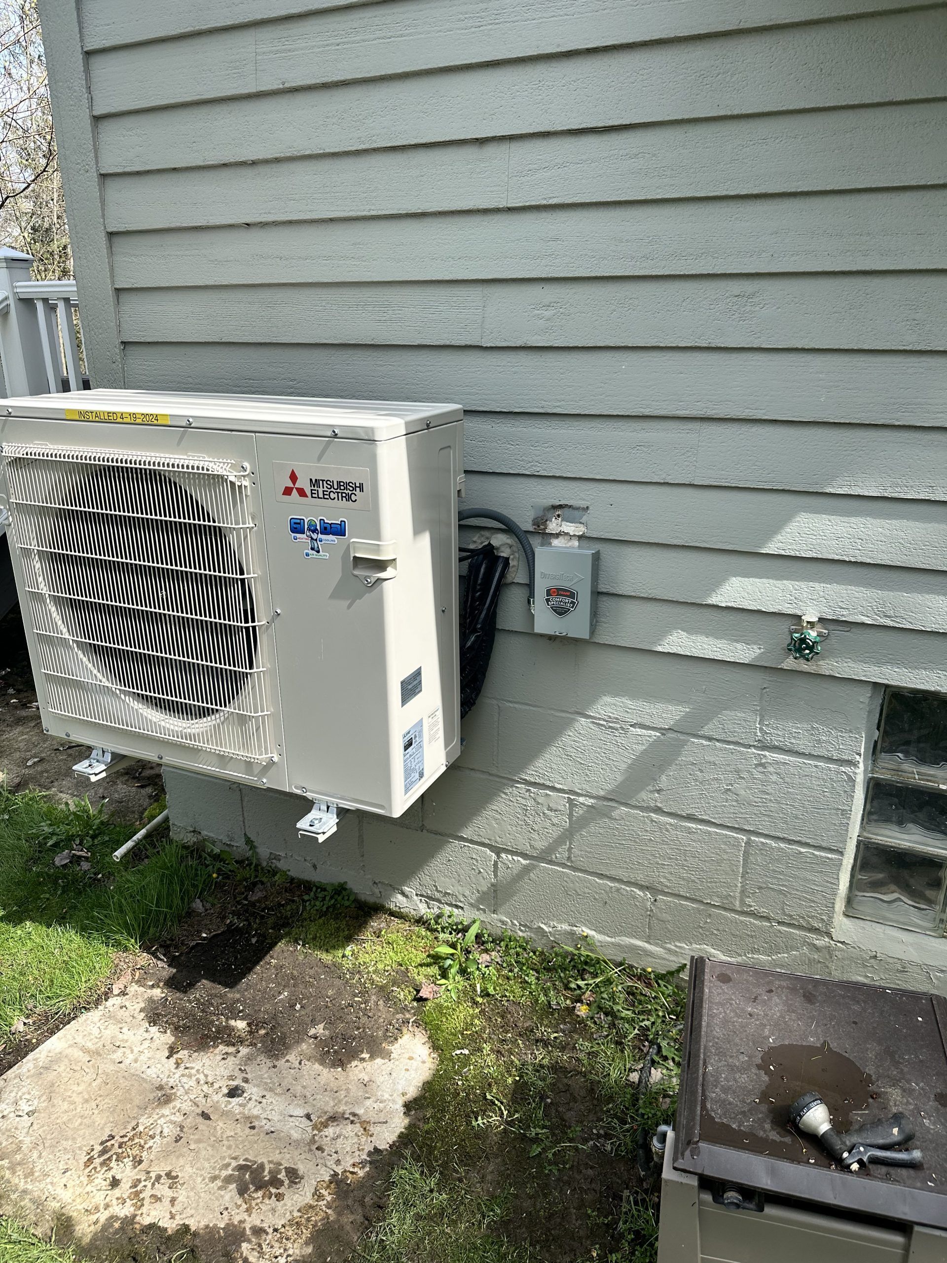 Mitsubishi air conditioner mounted on a gray brick foundation next to a gray wooden building.