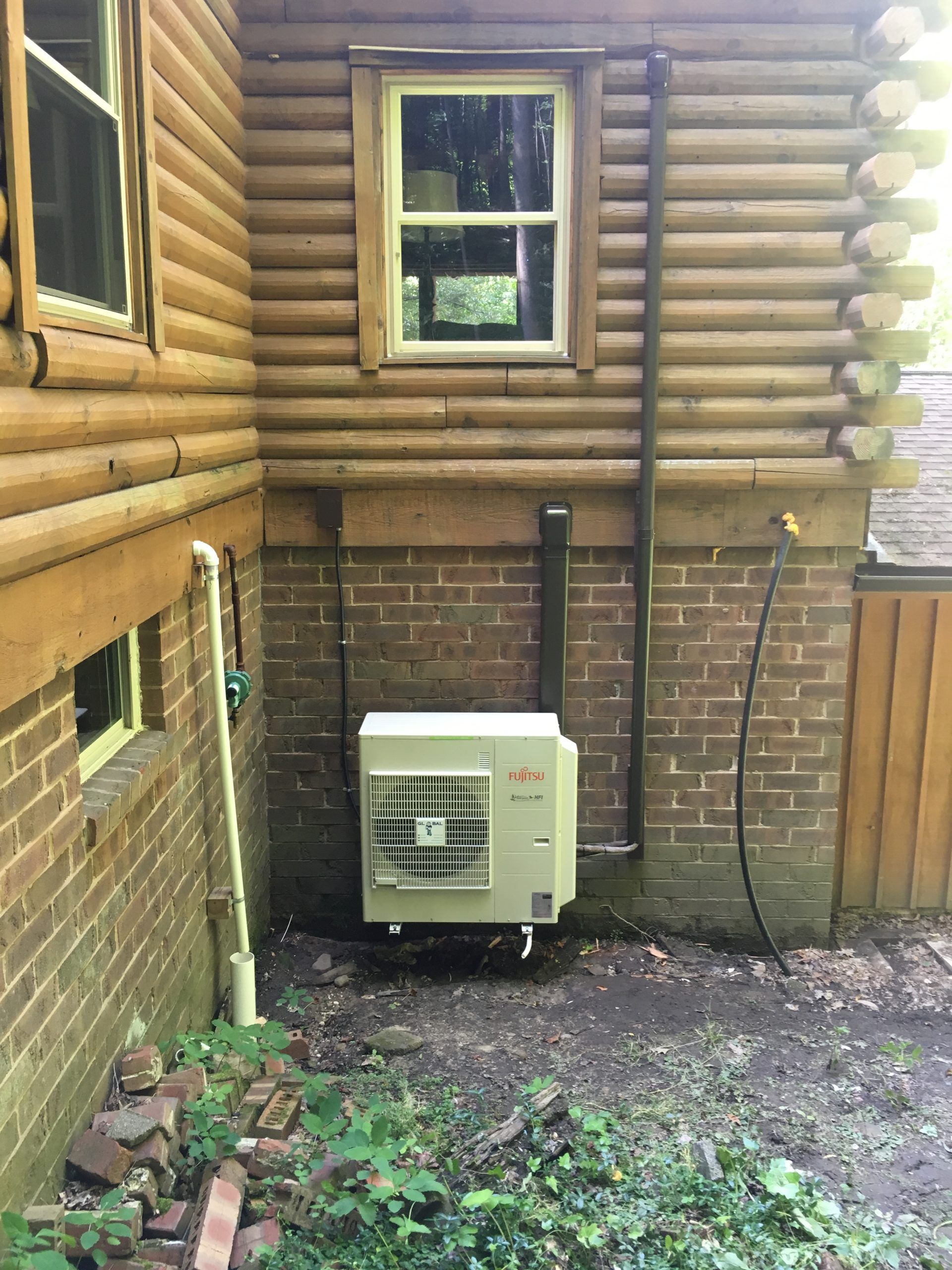 An exterior heat pump unit mounted on a brick foundation next to a log cabin.