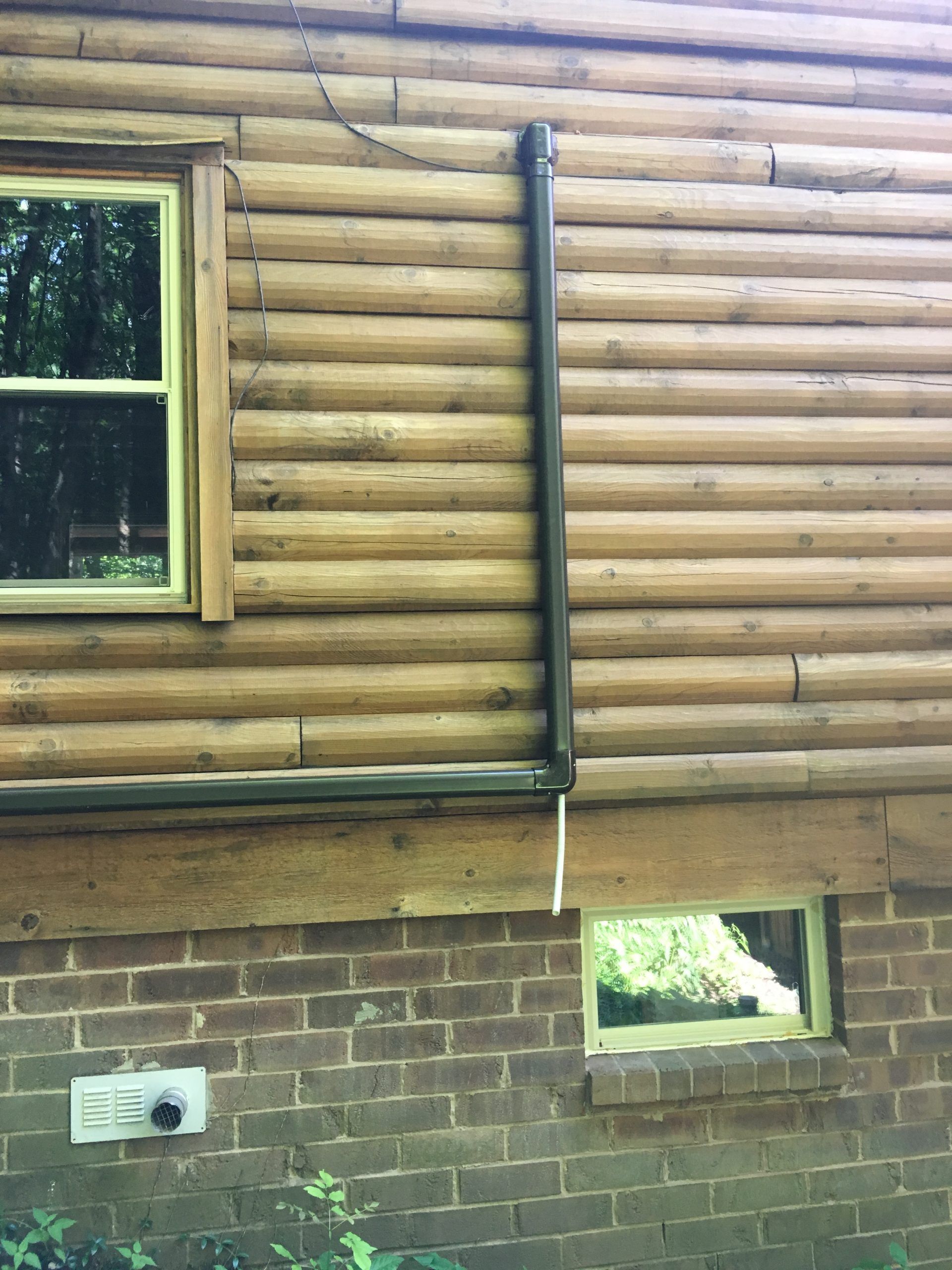 Exterior of a log cabin with a window, downspout, and a small basement window on a brick foundation.