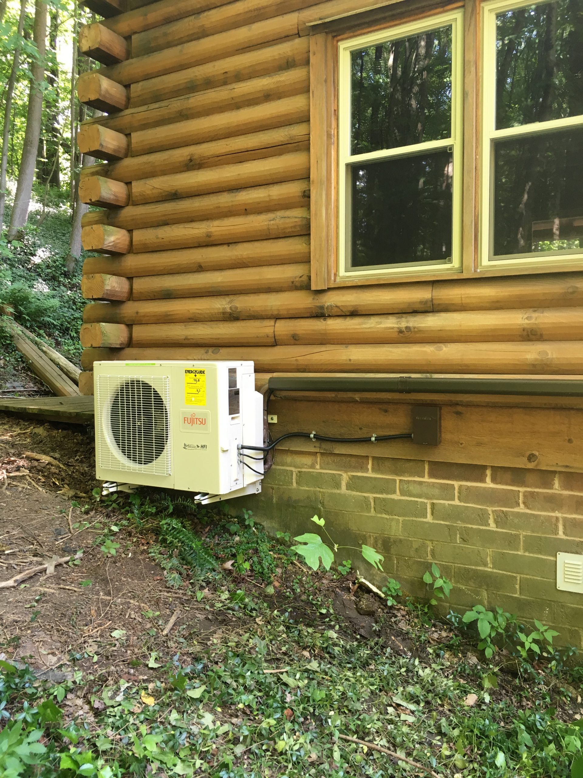 Exterior of a log cabin with an air conditioning unit attached to the wall near a window, set in a wooded area.