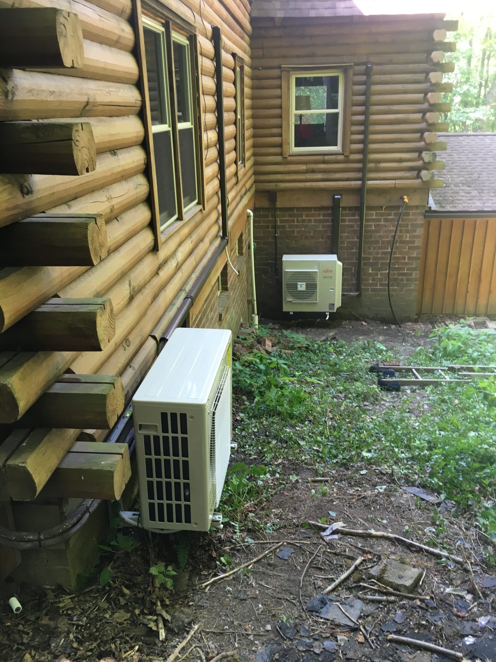 Two exterior air conditioning units mounted on a log cabin wall, with vegetation at their base.