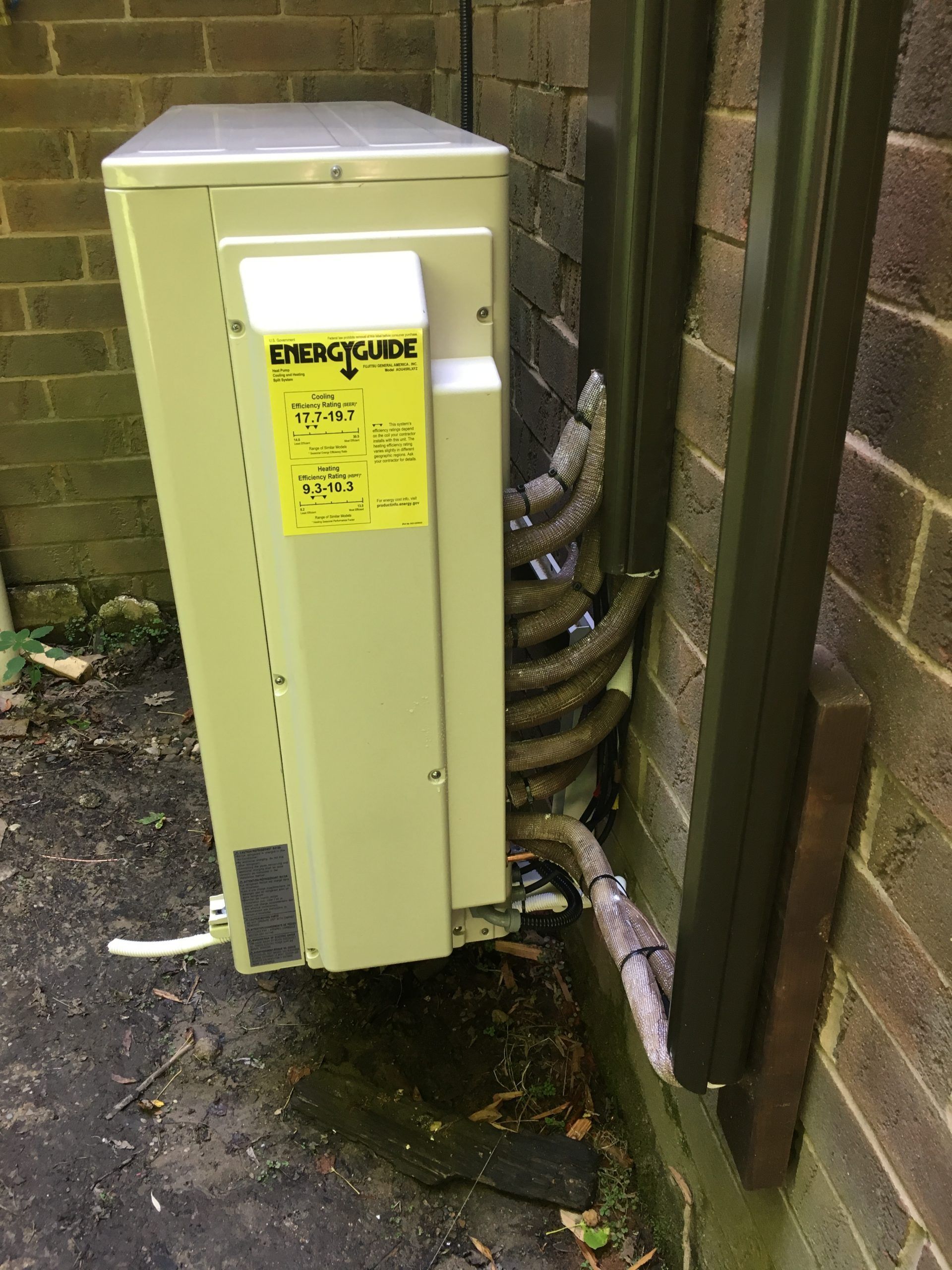 An outdoor air conditioning unit with connected piping, mounted against a brick wall. An energy guide sticker is visible.