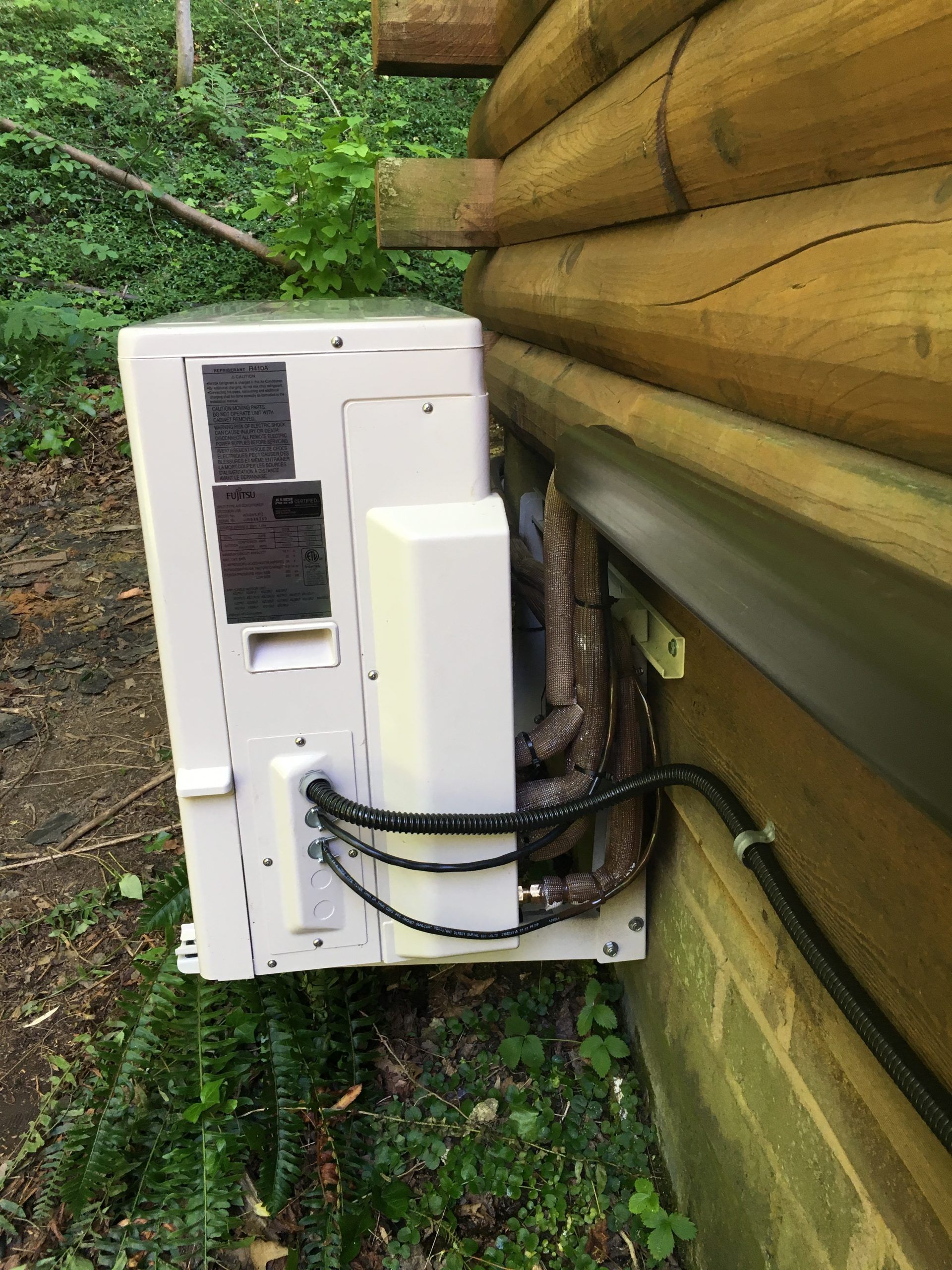 Outdoor HVAC unit mounted on a wooden cabin wall, with black tubing and a metal bracket.
