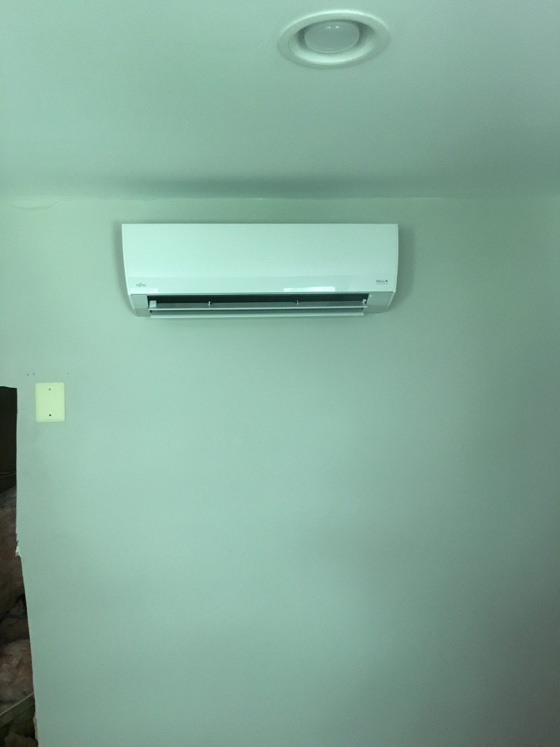 White wall-mounted air conditioner on a pale green wall, below a ceiling light.