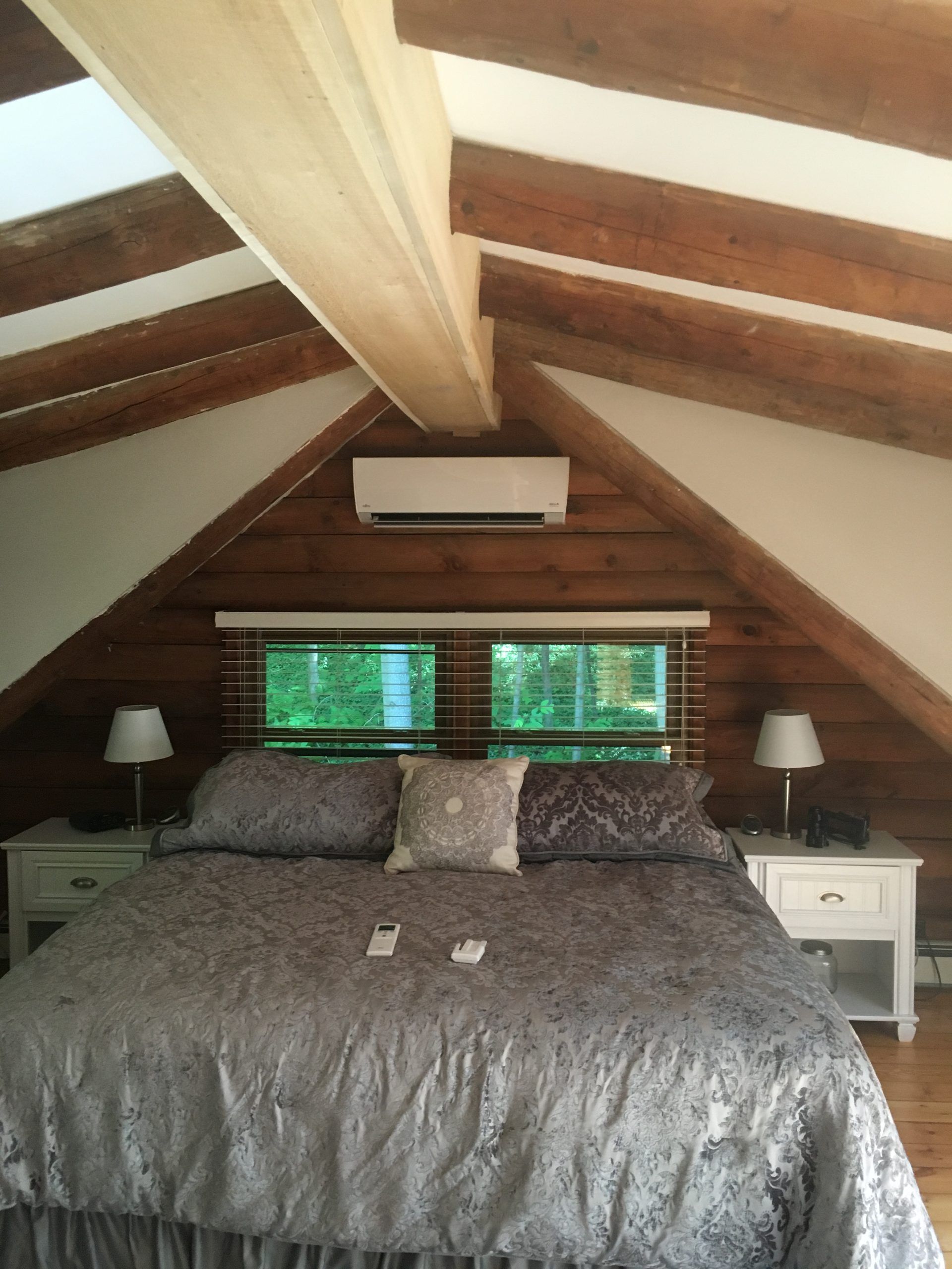 Bedroom with bed, nightstands, and air conditioning unit against log-paneled wall, beams overhead.
