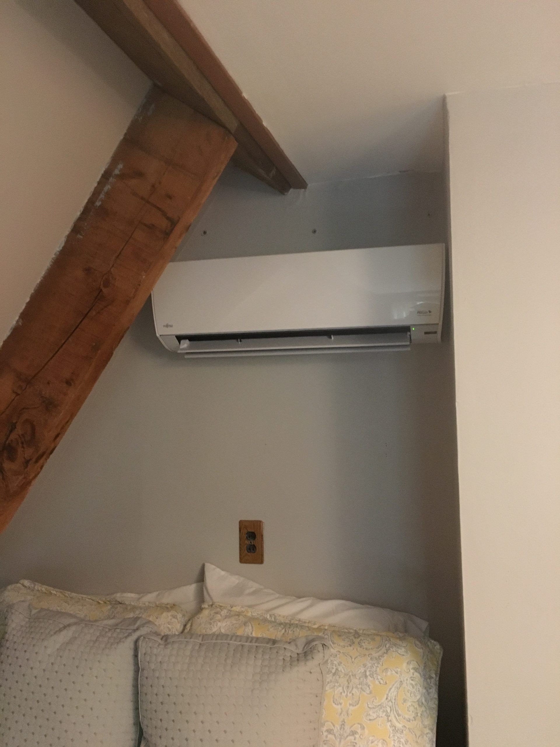 White air conditioner on gray wall near wooden beam and bed with pillows.