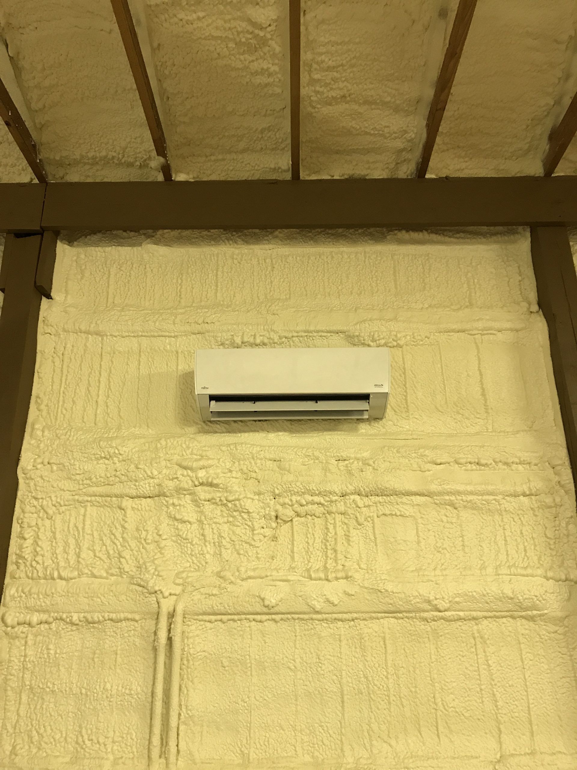 White air conditioning unit mounted on a foam-insulated wall, under a wood-beamed ceiling.