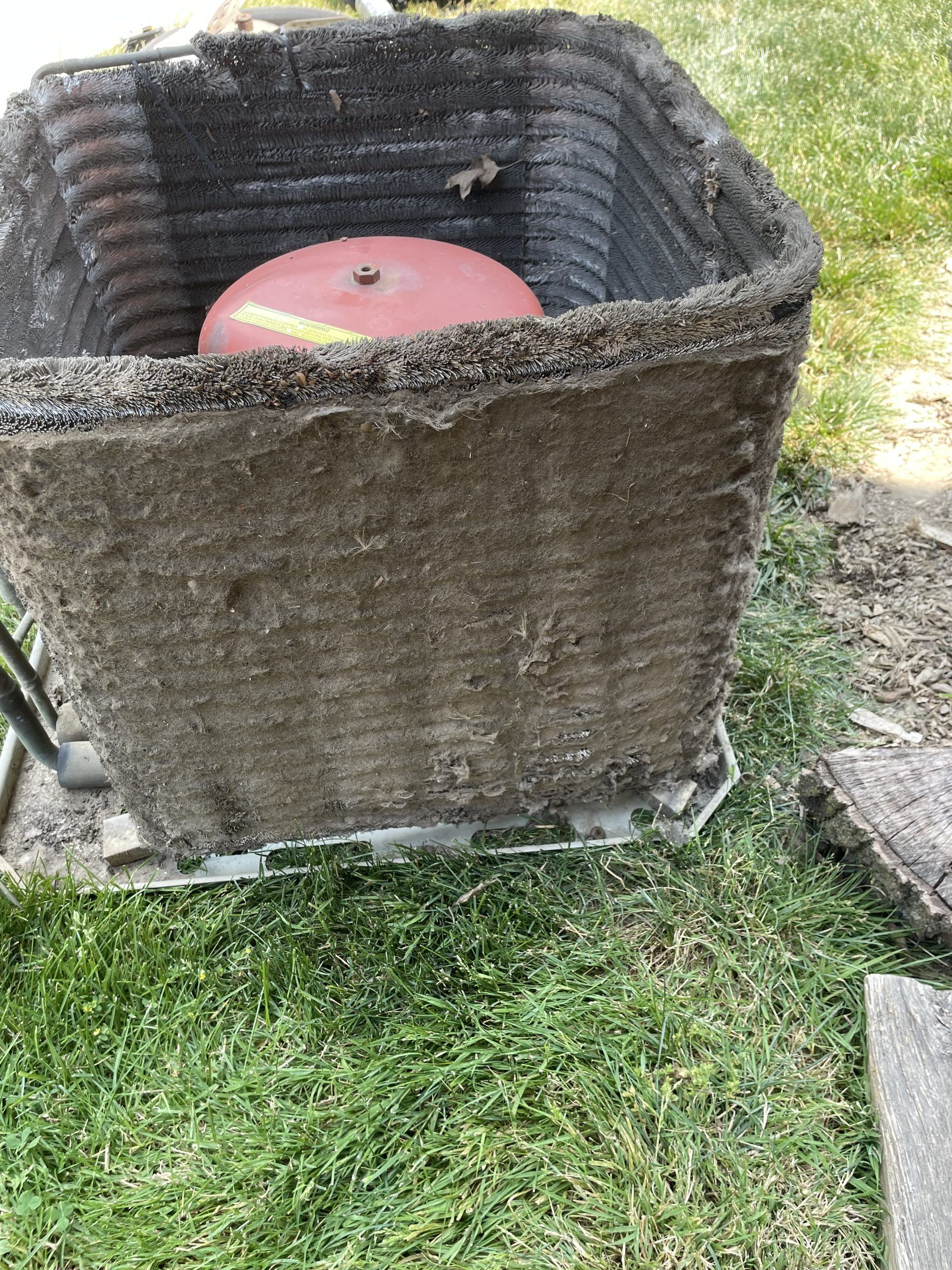 Outdoor air conditioning unit with a red tank inside. Dark gray, concrete-like exterior with a green grassy lawn.