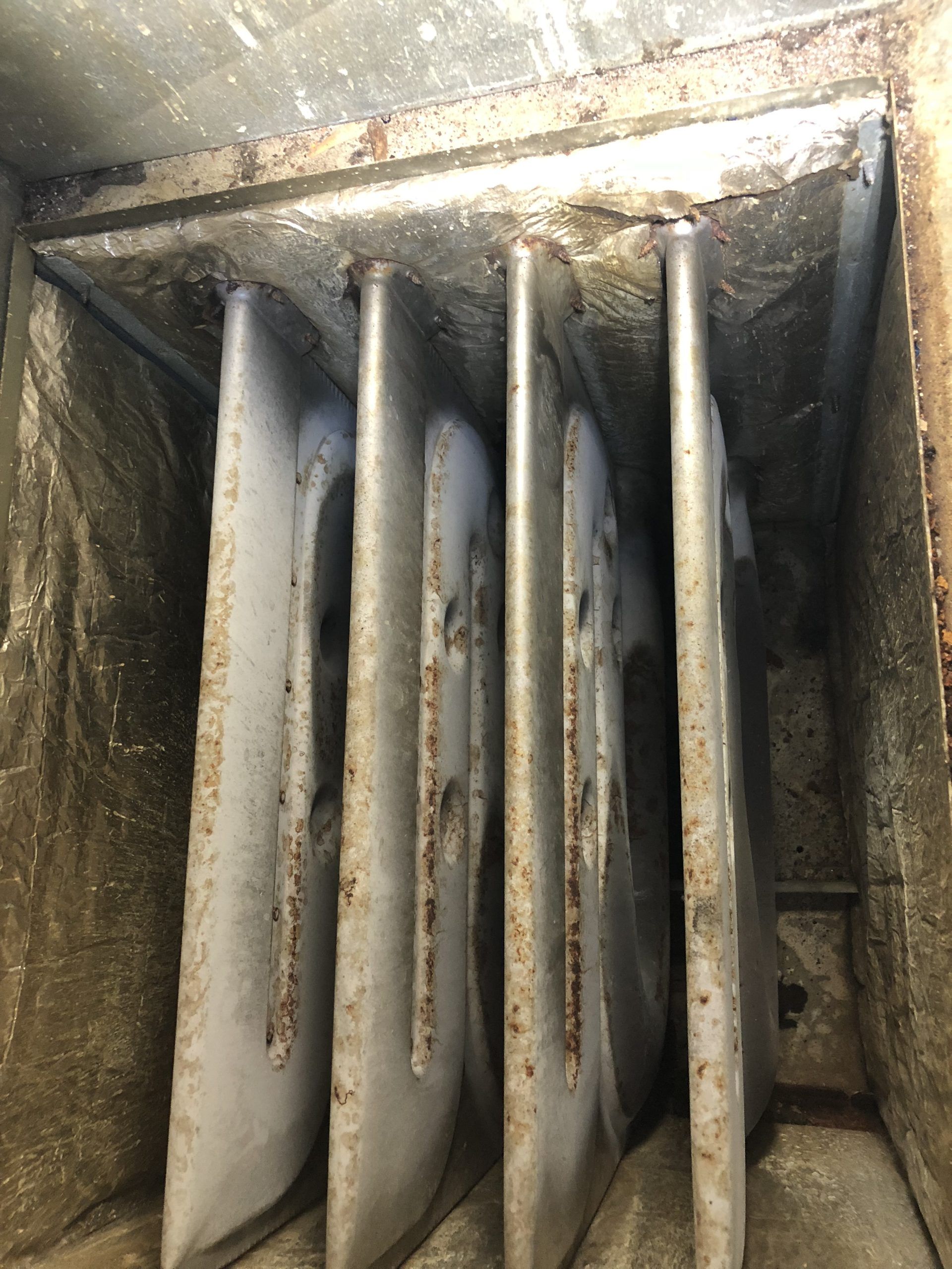 Inside a dusty rectangular metal air duct are five vertical, curved metal vanes.