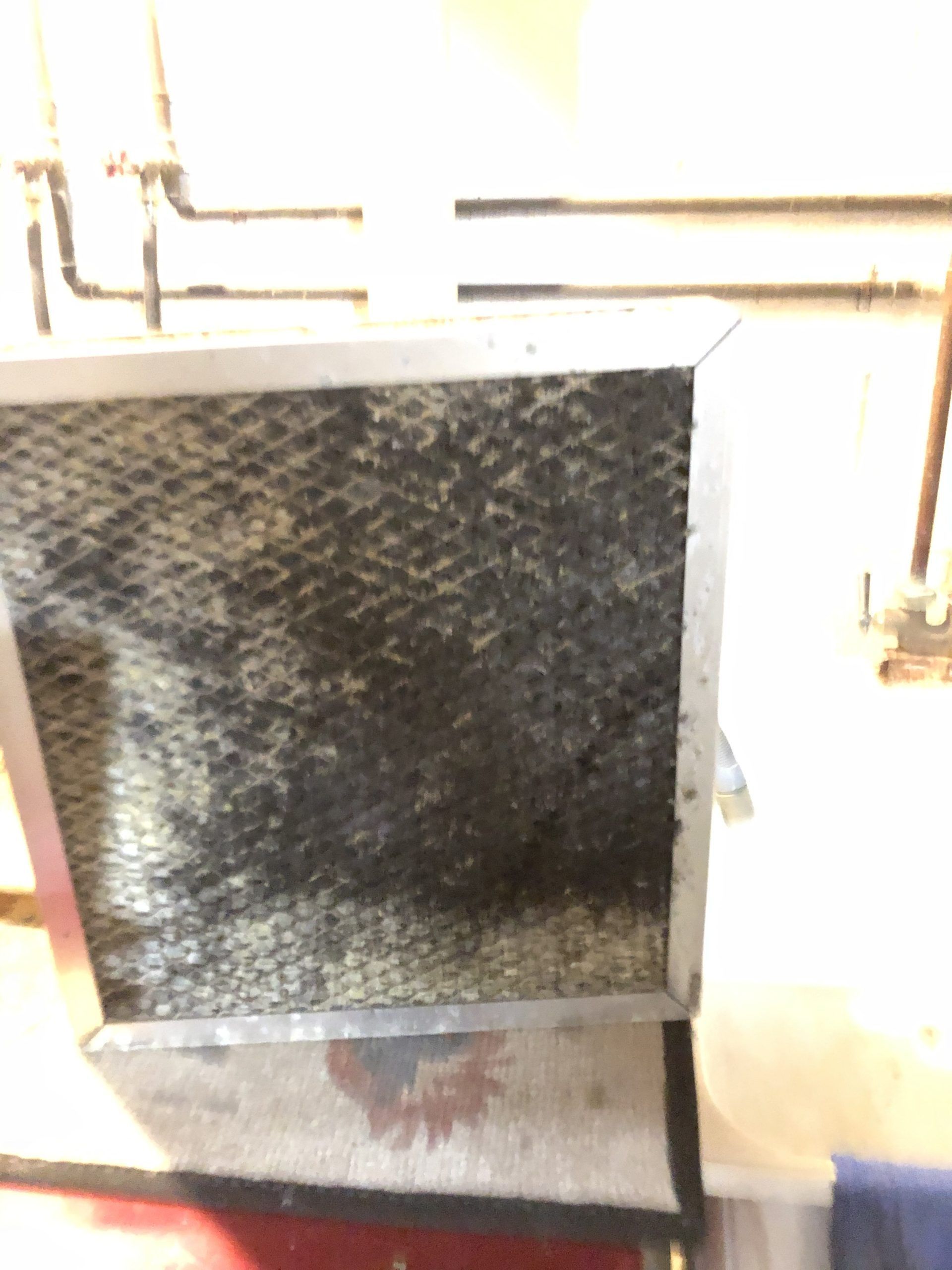 Dirty, black, clogged air filter in a metal frame, mounted near pipes.