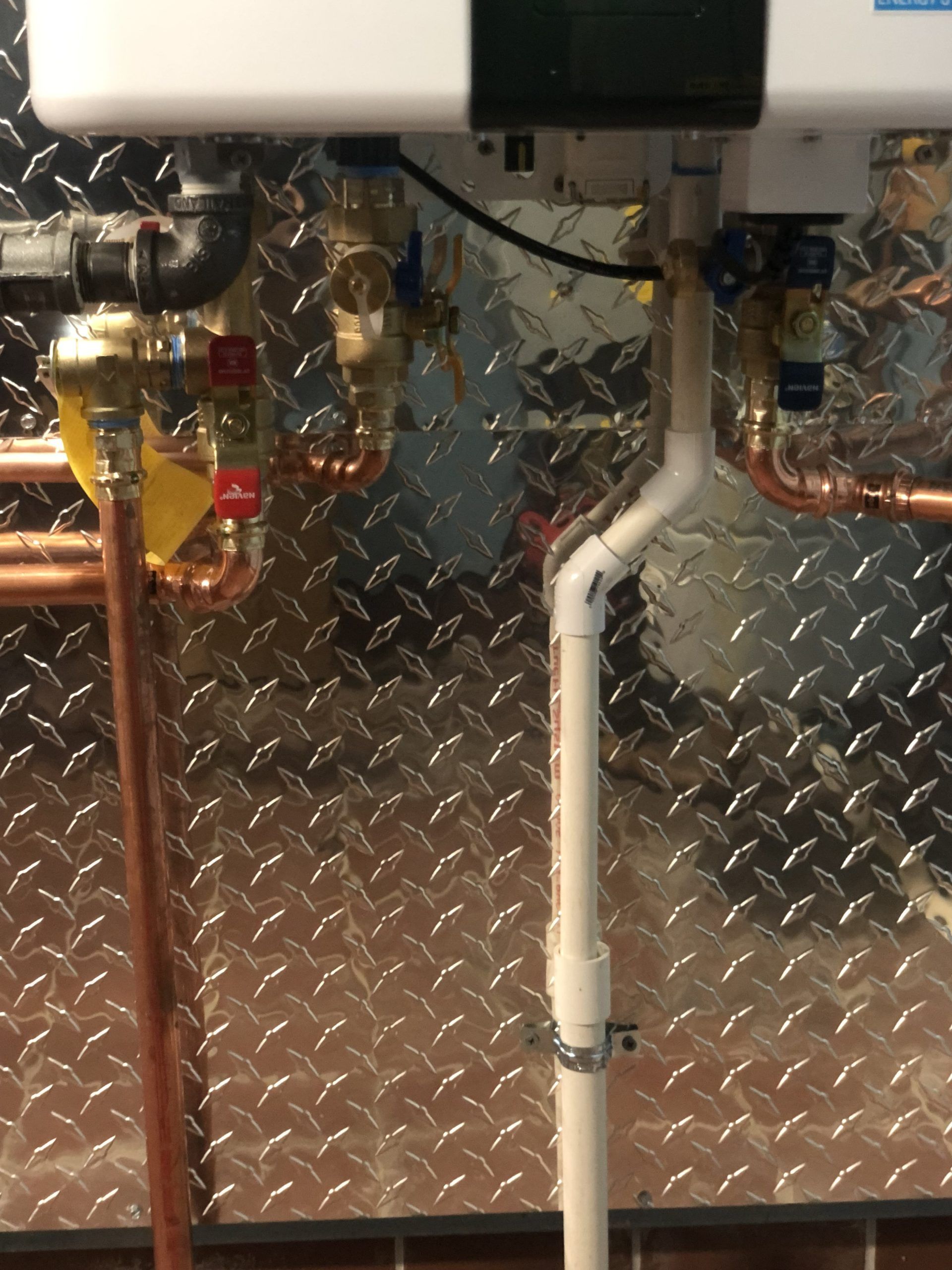 Plumbing: a white water heater with copper and PVC pipes and valves against a diamond-plate wall.