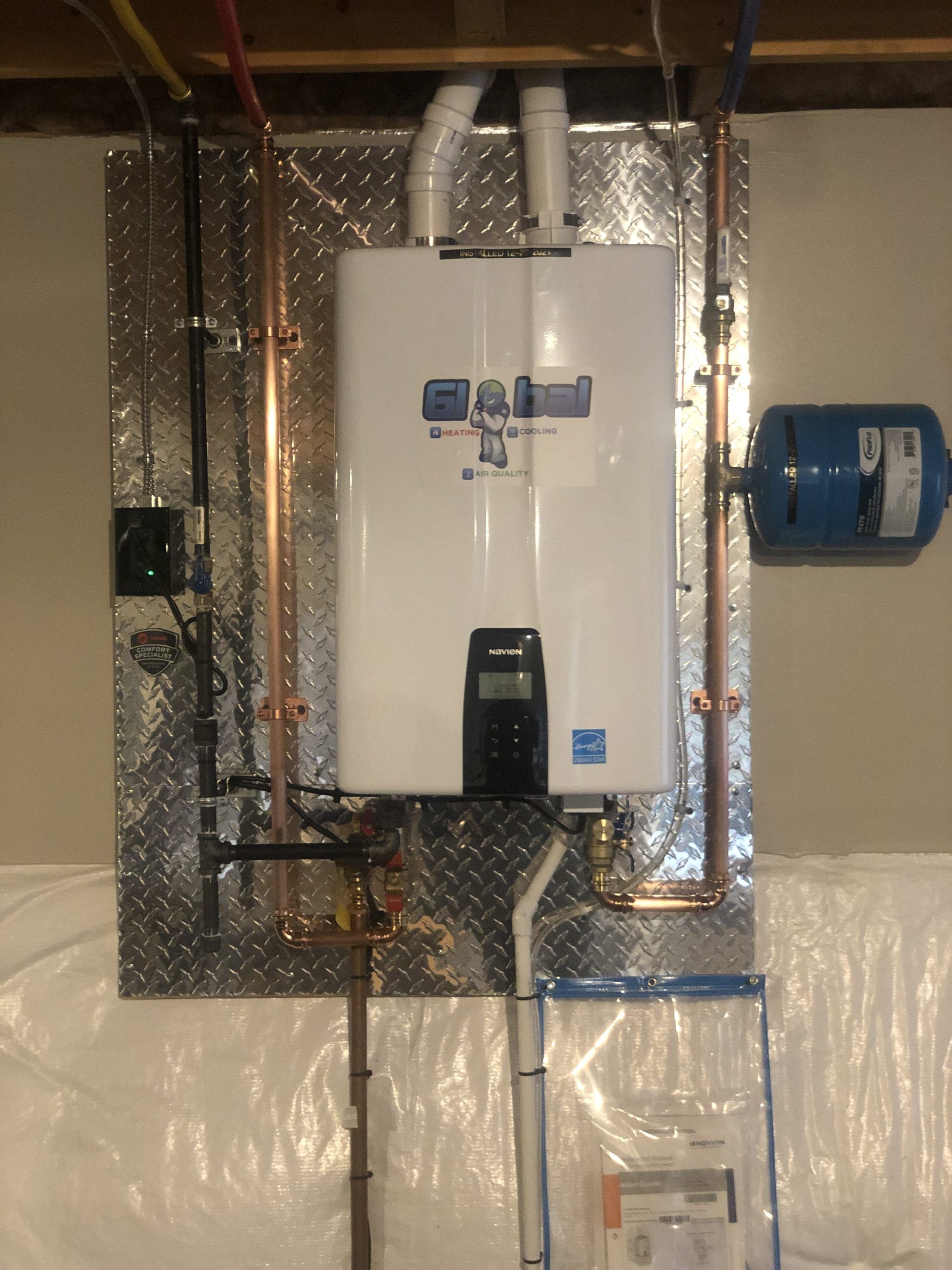 Tankless water heater mounted on a metal plate in a basement, with copper pipes and a blue expansion tank.