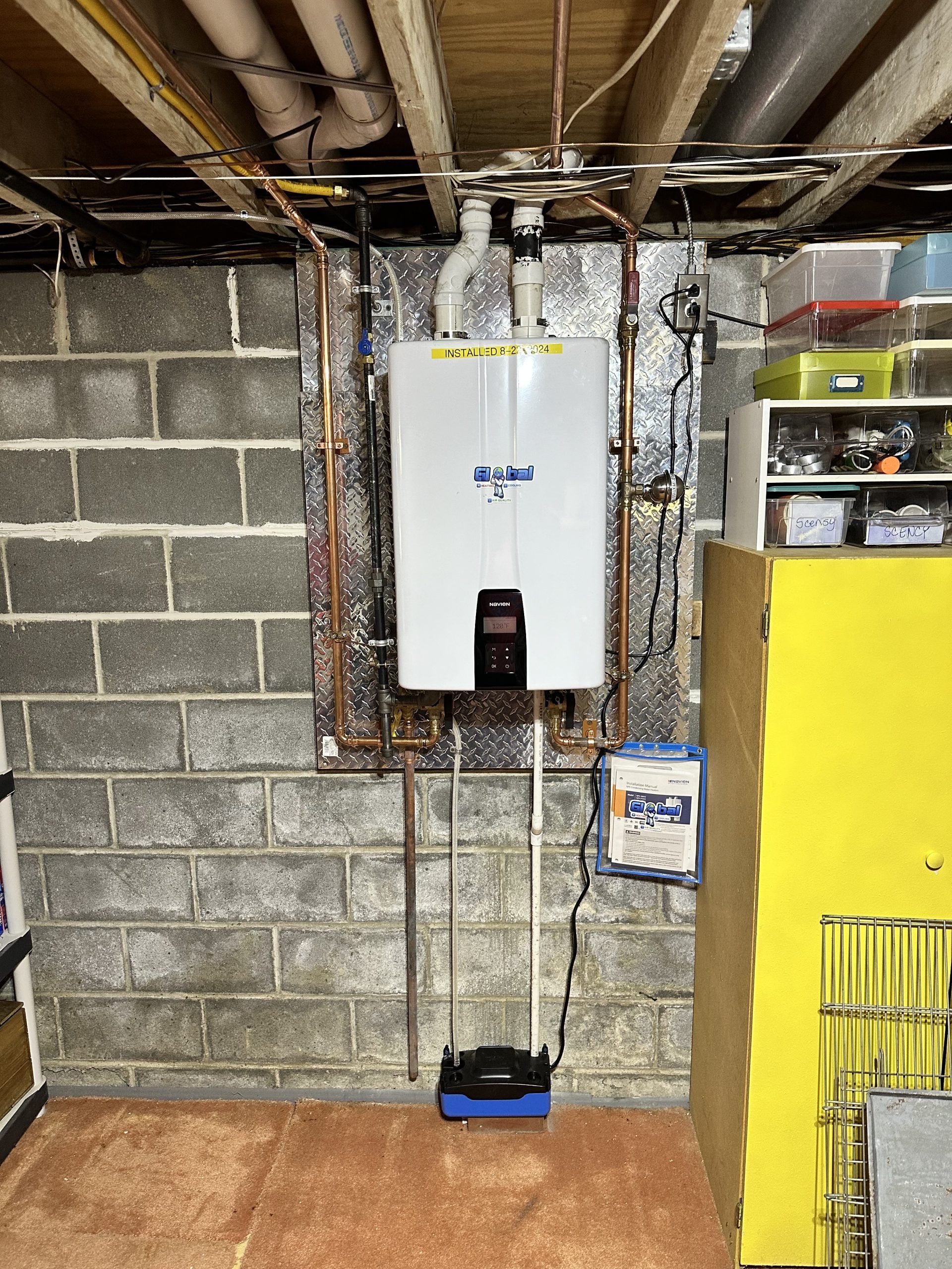 A wall-mounted white water heater in a basement with exposed pipes and a concrete block wall.