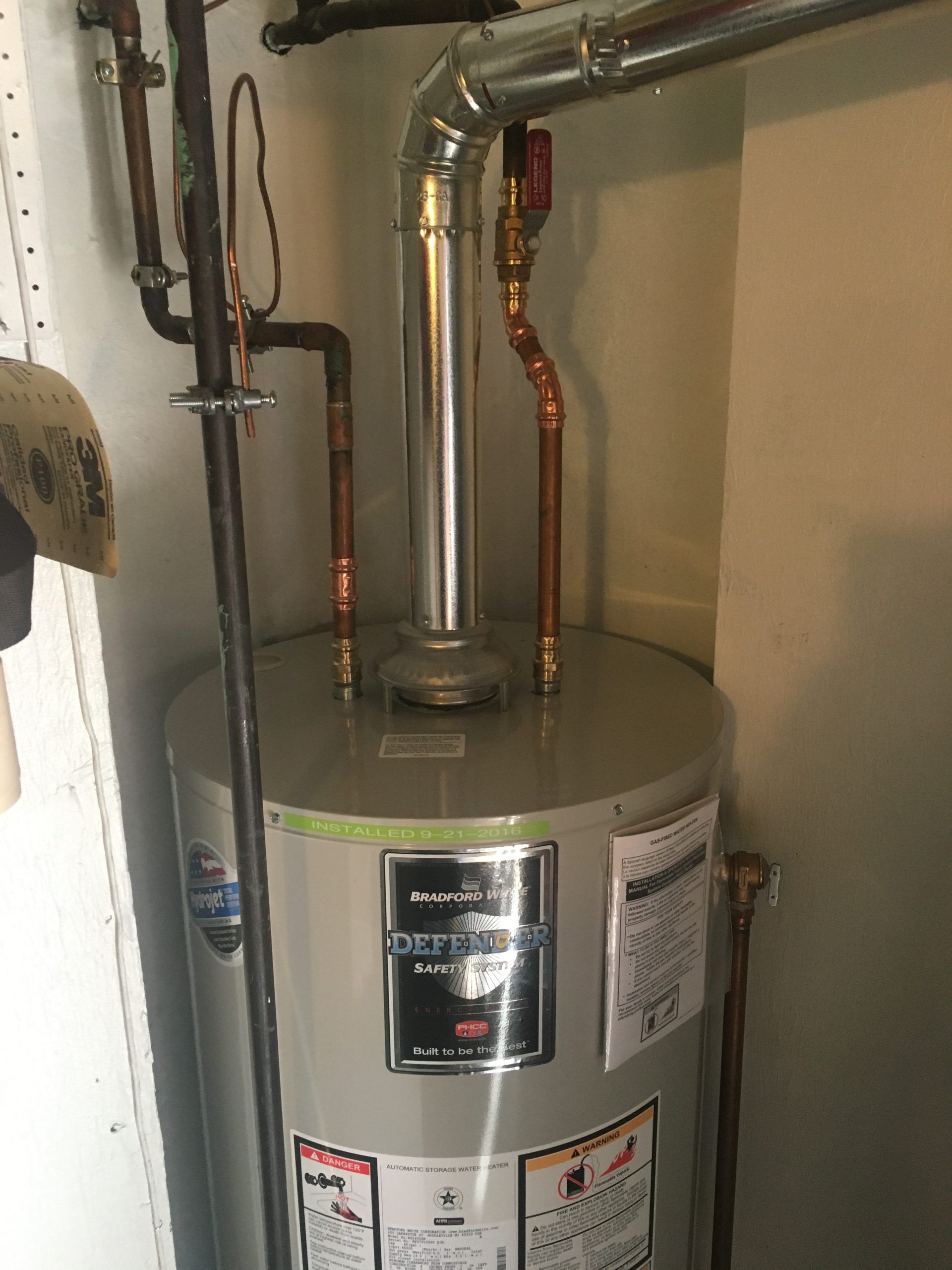 Water heater in a utility closet with copper pipes, a metal vent, and warning labels.