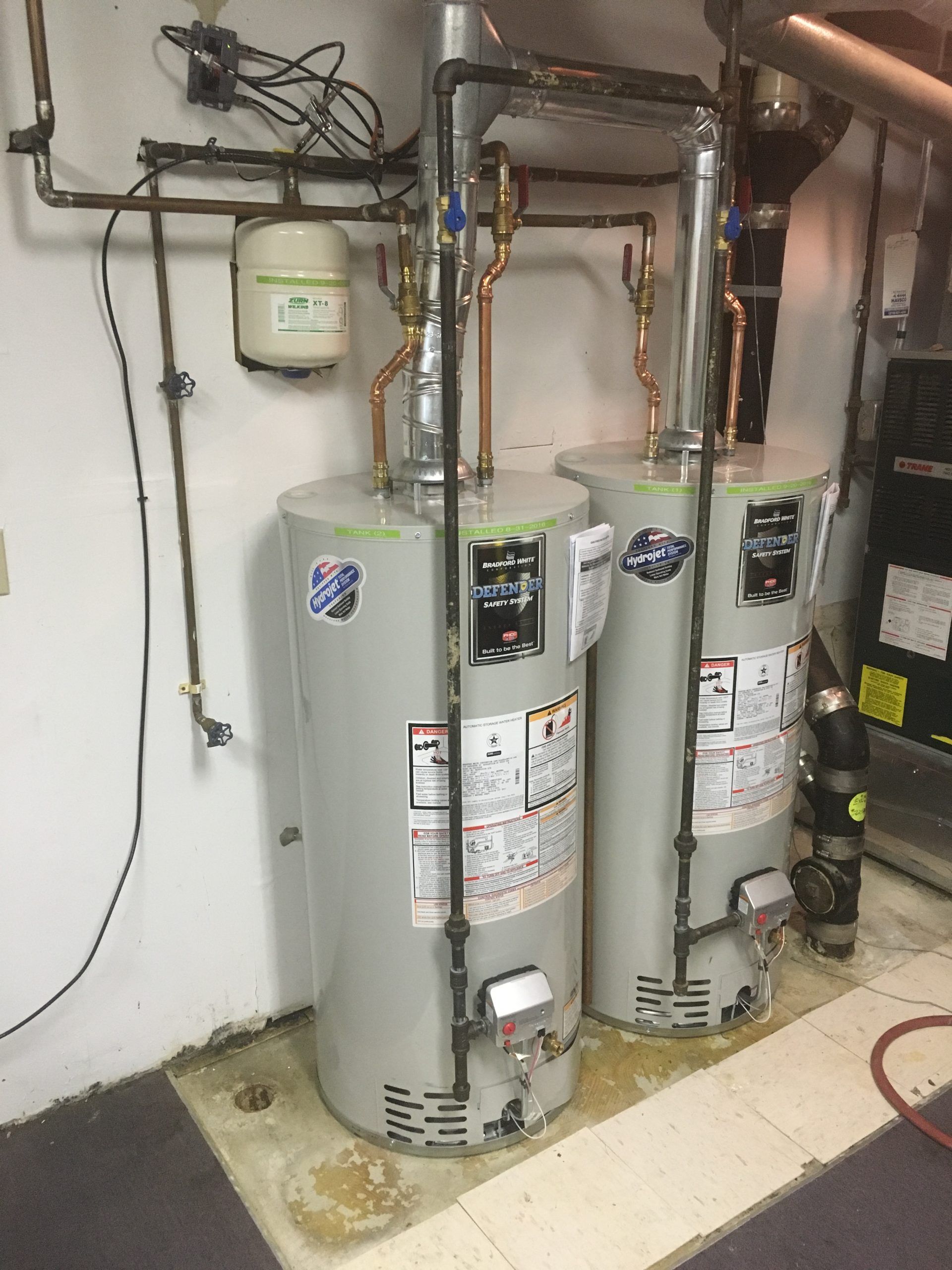 Two water heaters with copper pipes in a basement. A white expansion tank is mounted on the wall.