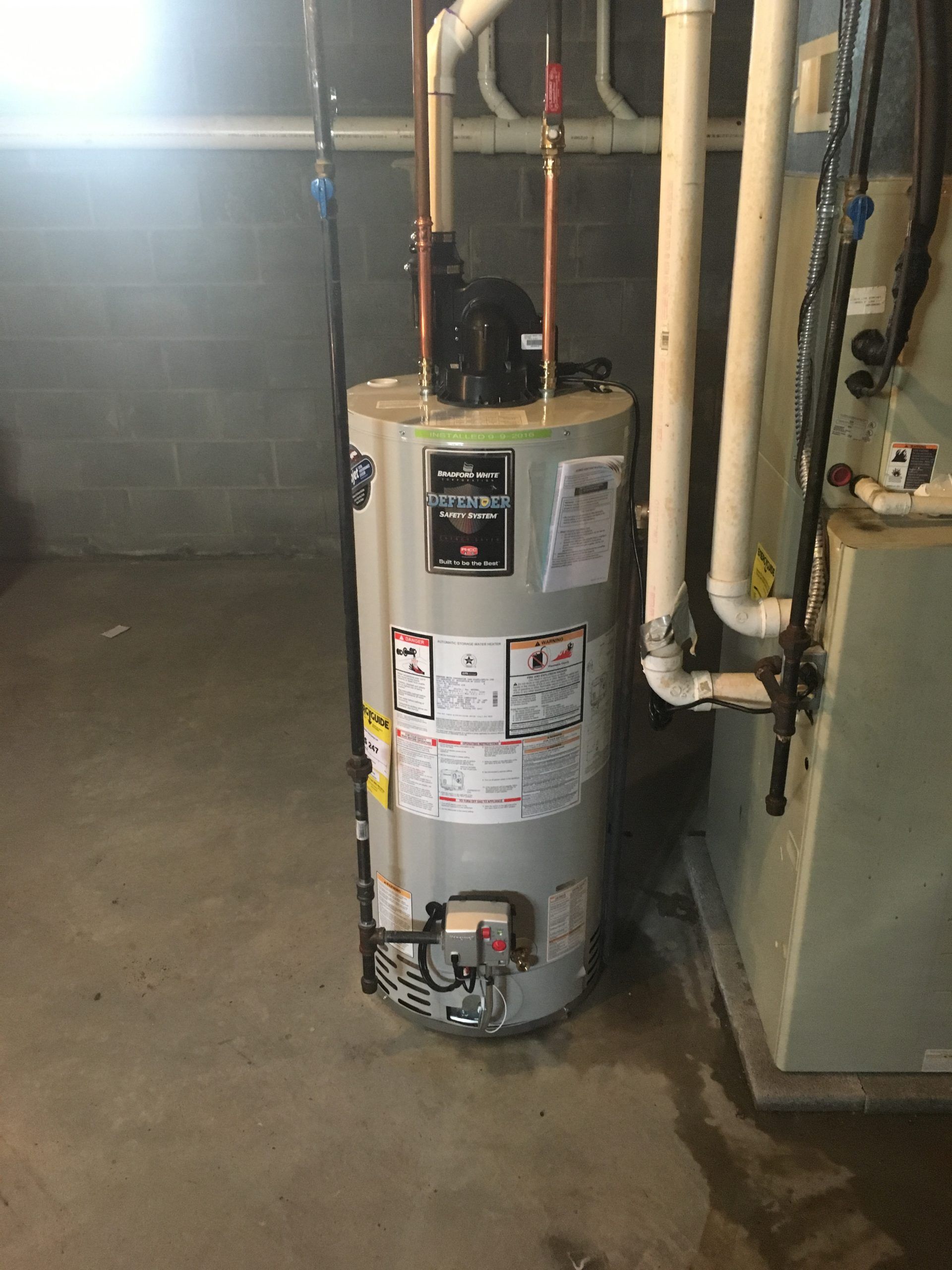 Water heater in a basement.  Gray tank with pipes and a furnace to the right. Dark concrete floor.
