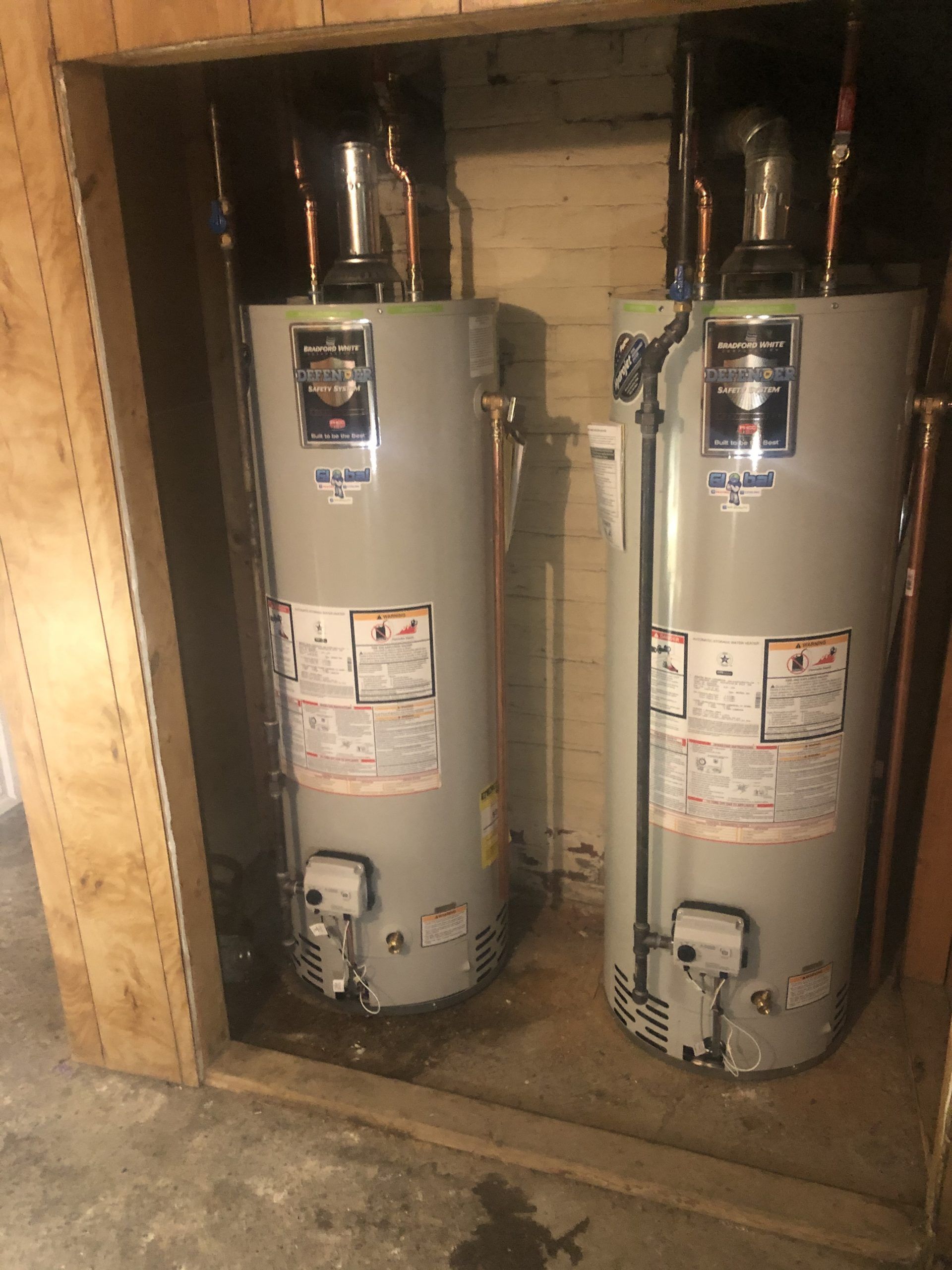 Two water heaters in a wood-lined alcove, with silver tanks, pipes, and labels.