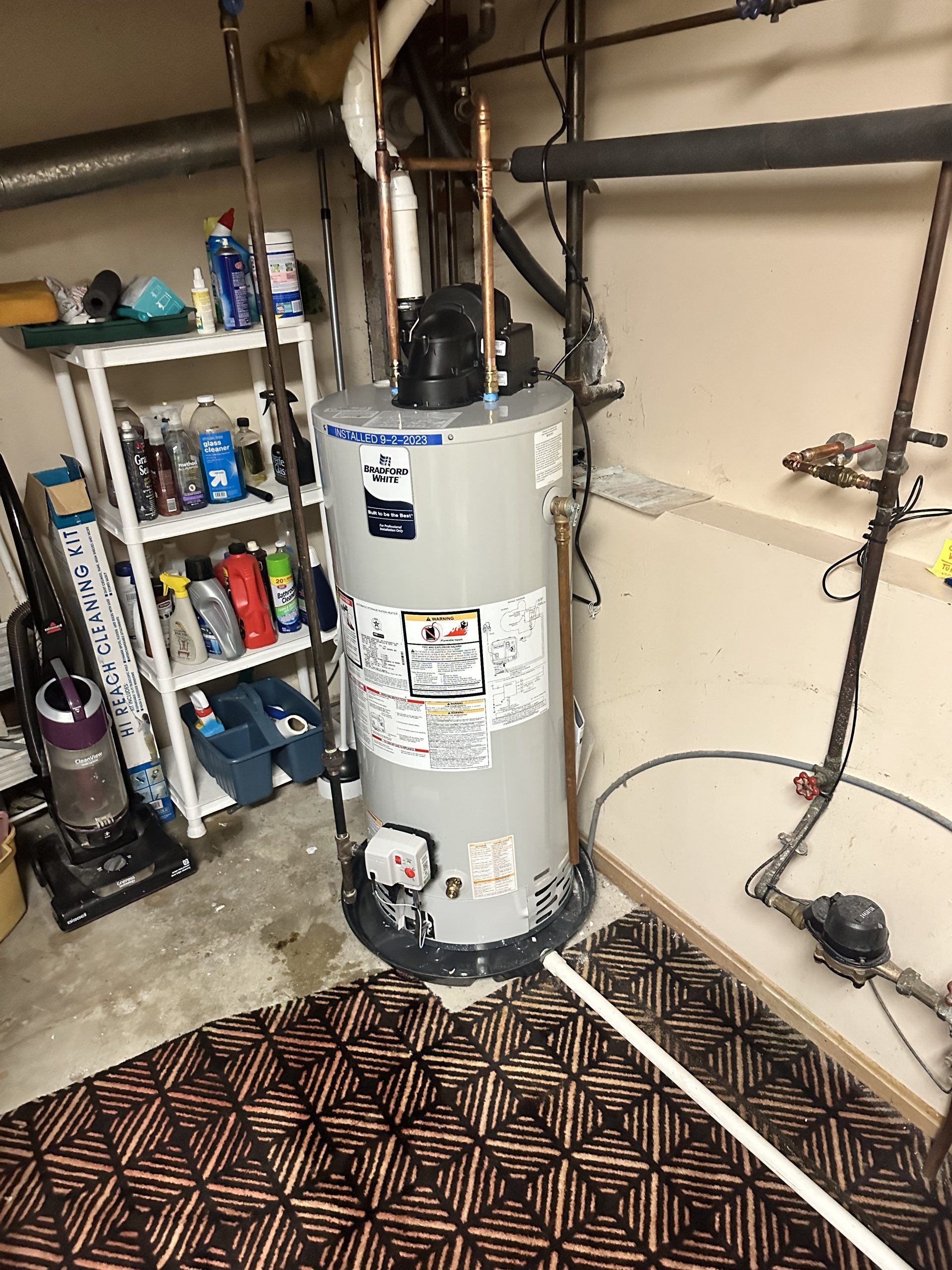 Water heater in a garage setting with pipes and shelving nearby.