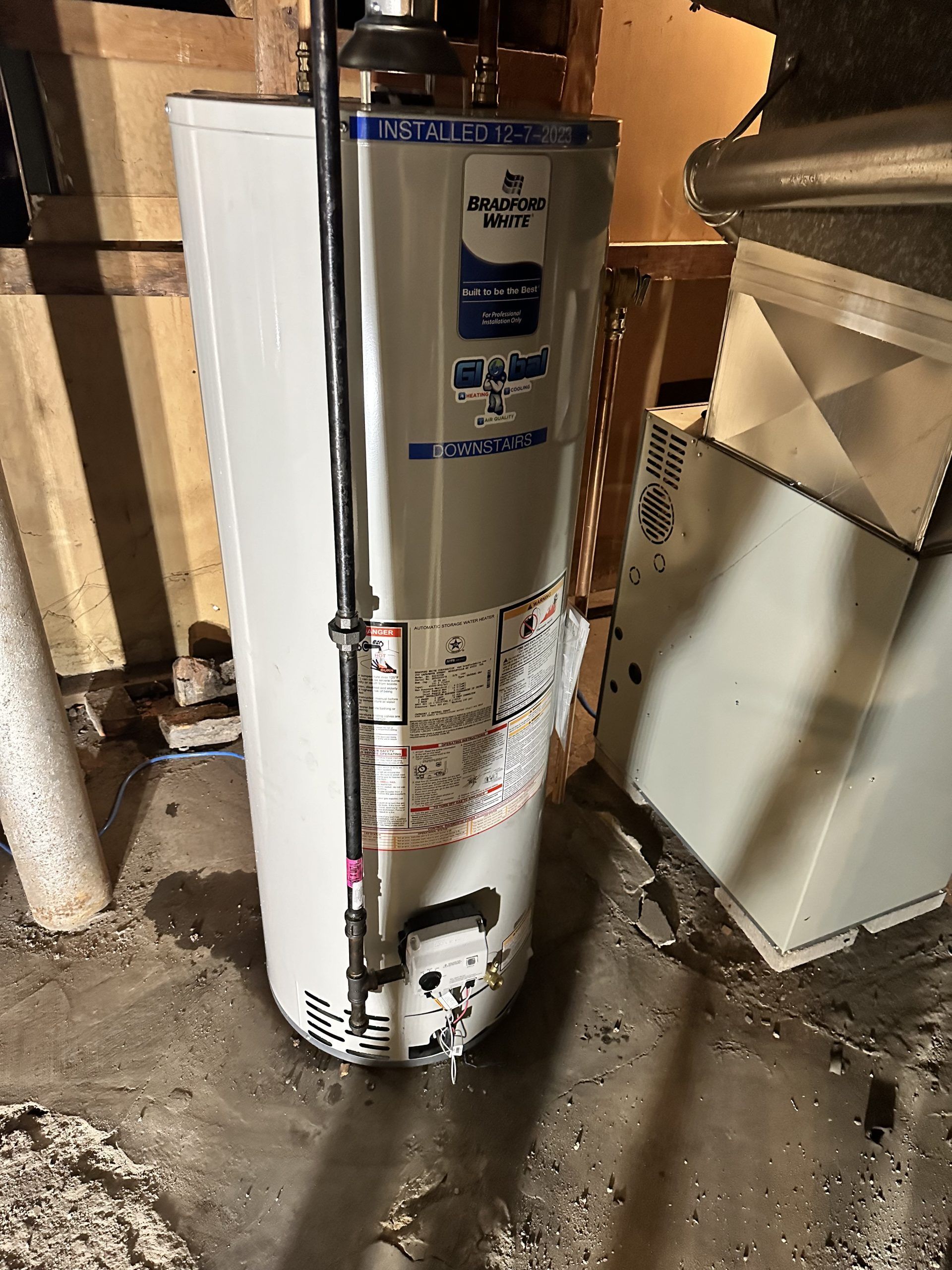 A water heater in a basement with water on the floor. It is white and blue.