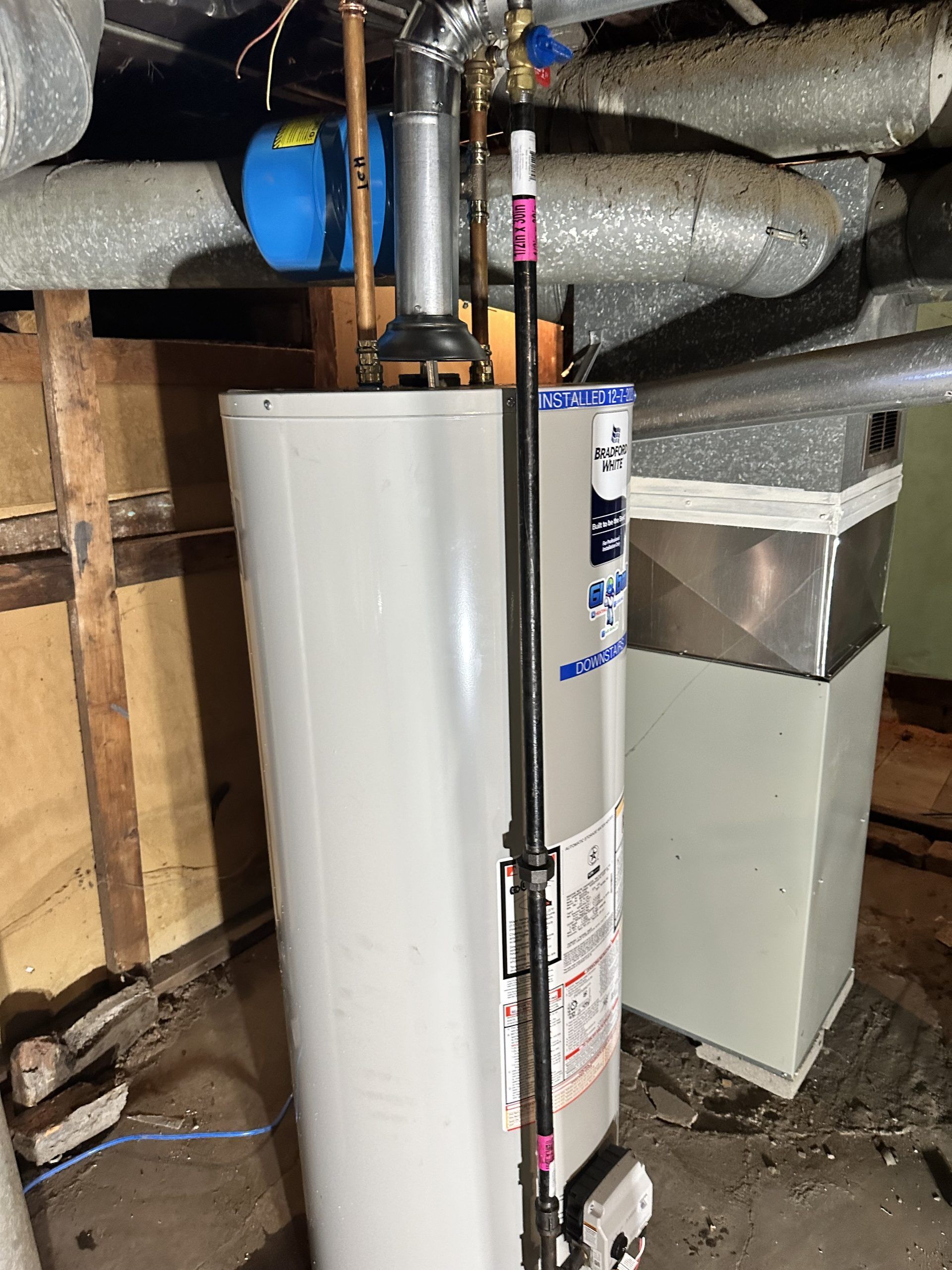 Water heater in a basement.  Silver tank with vent pipe, black pipe running from top to bottom.