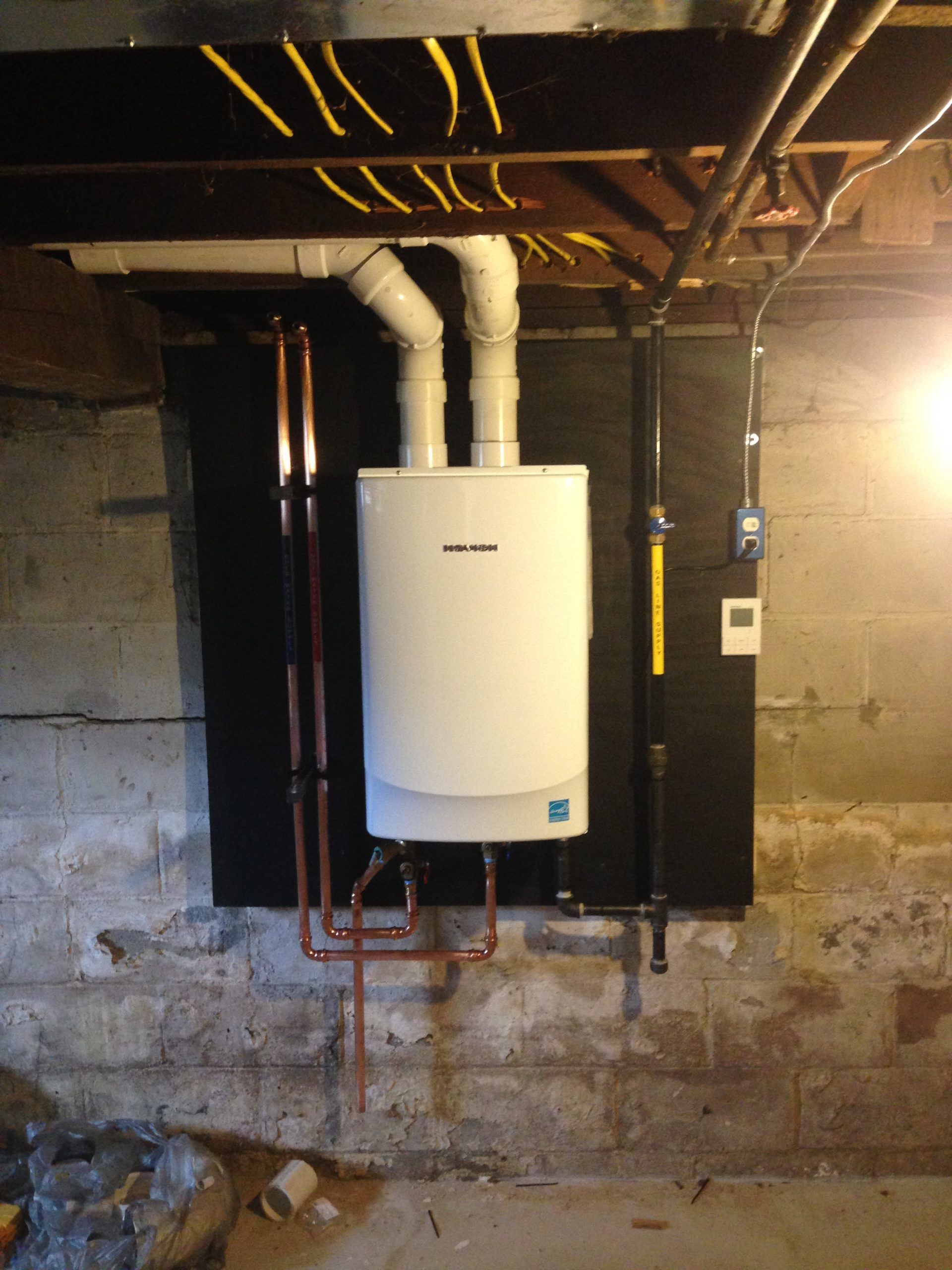 White wall-mounted boiler with copper and black pipes in a basement.