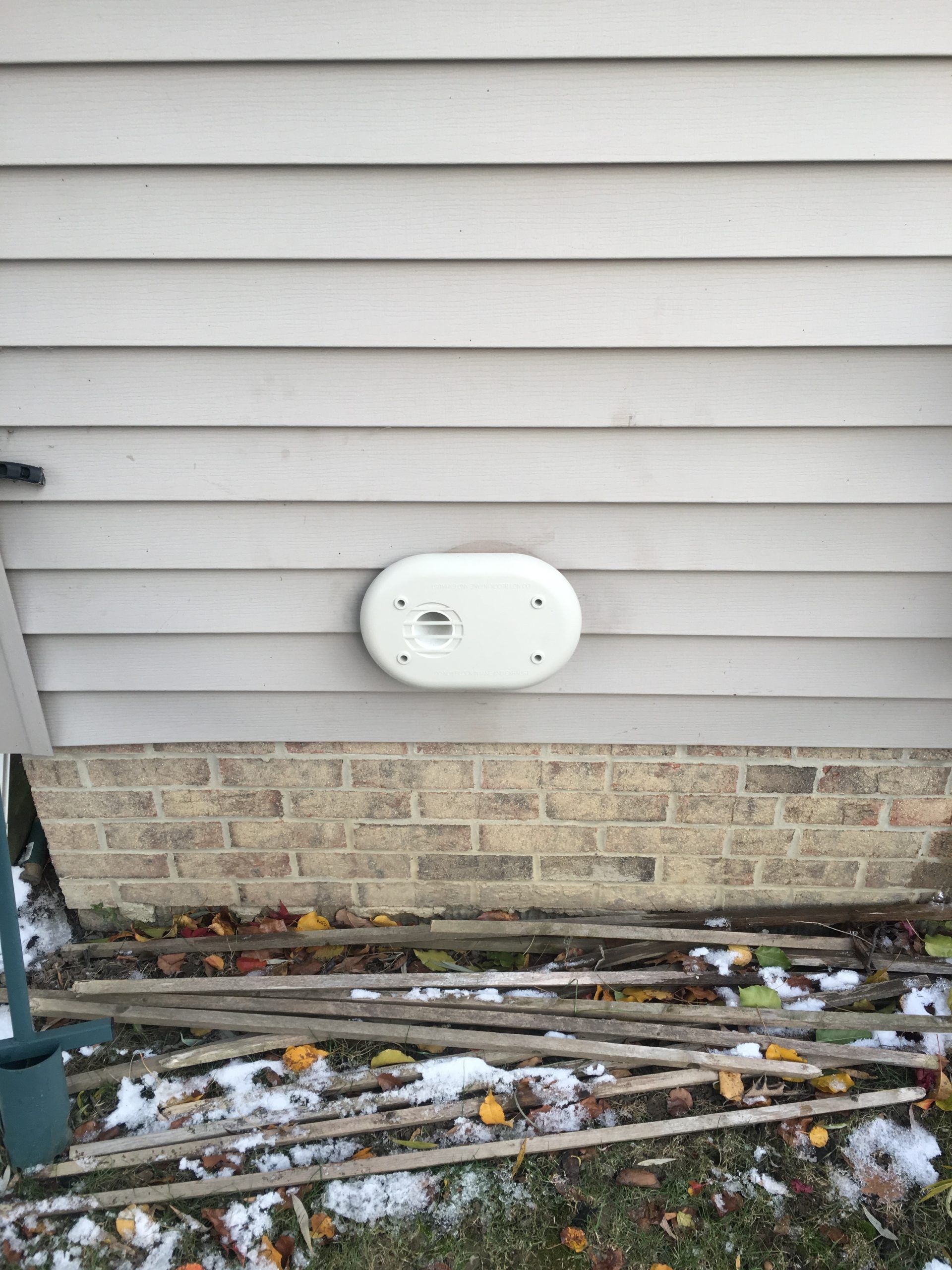White oval vent on a gray siding house wall above a brick foundation.