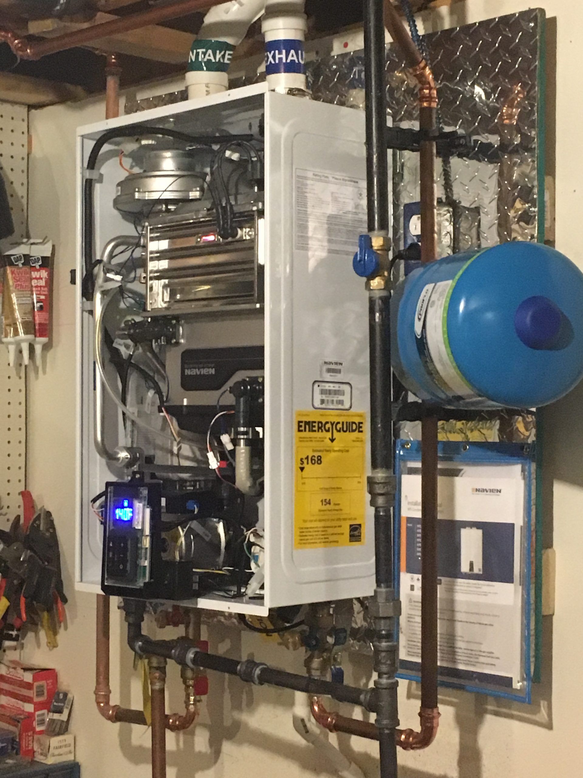Wall-mounted, open-faced boiler unit with copper pipes and a blue expansion tank on a beige wall.