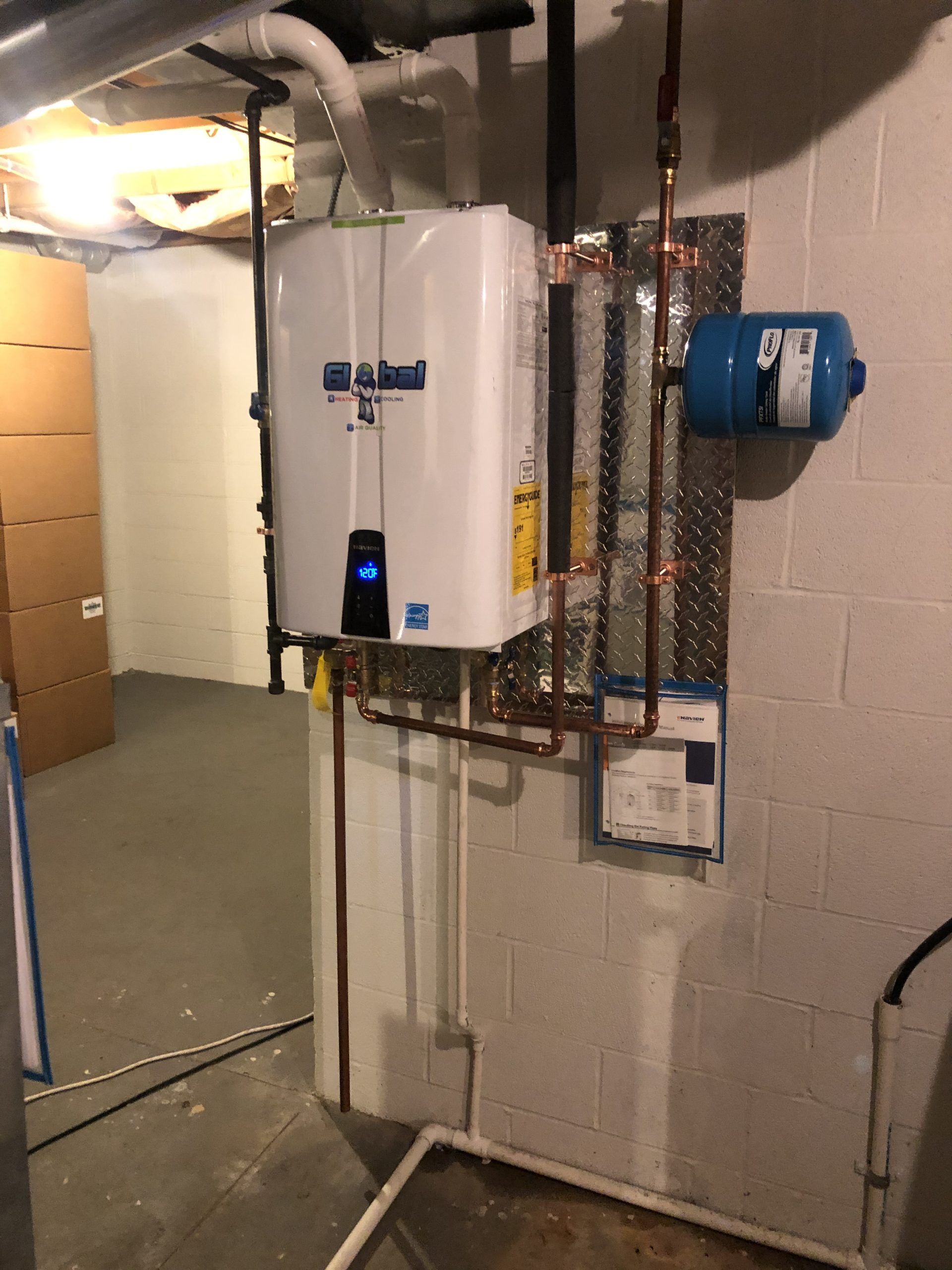 White tankless water heater with copper pipes, blue expansion tank, and electrical panel on a concrete wall.