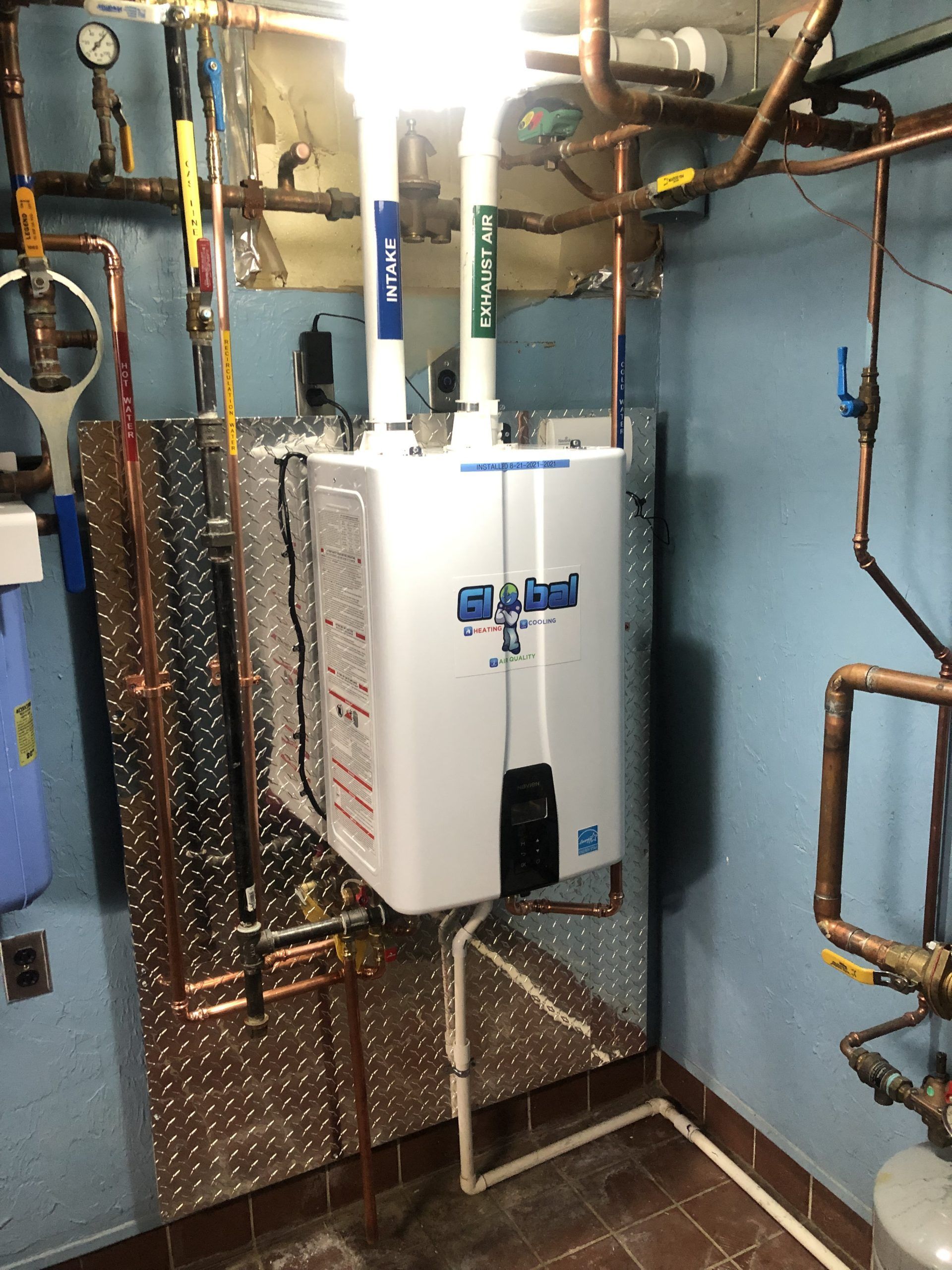 Tankless water heater in a utility room. Copper pipes, blue and green water lines, diamond plate backing.
