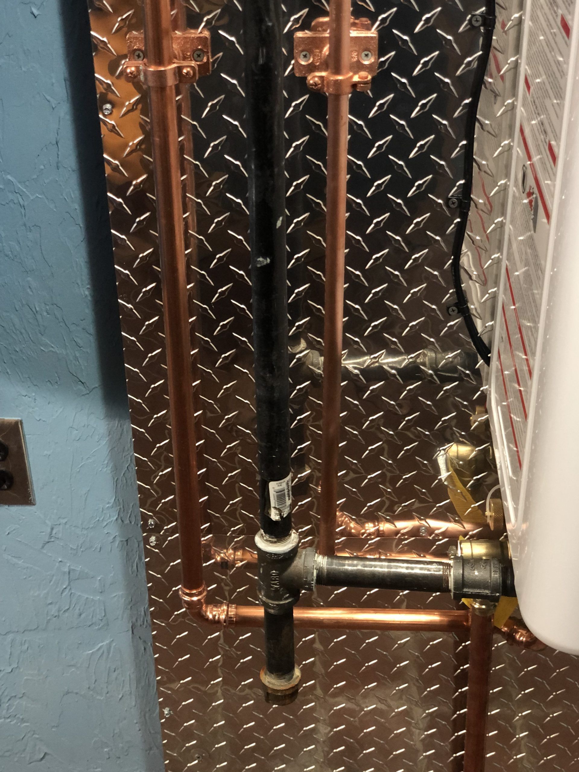 Copper pipes and black pipe against a diamond-plate metal panel, part of a water heater installation.