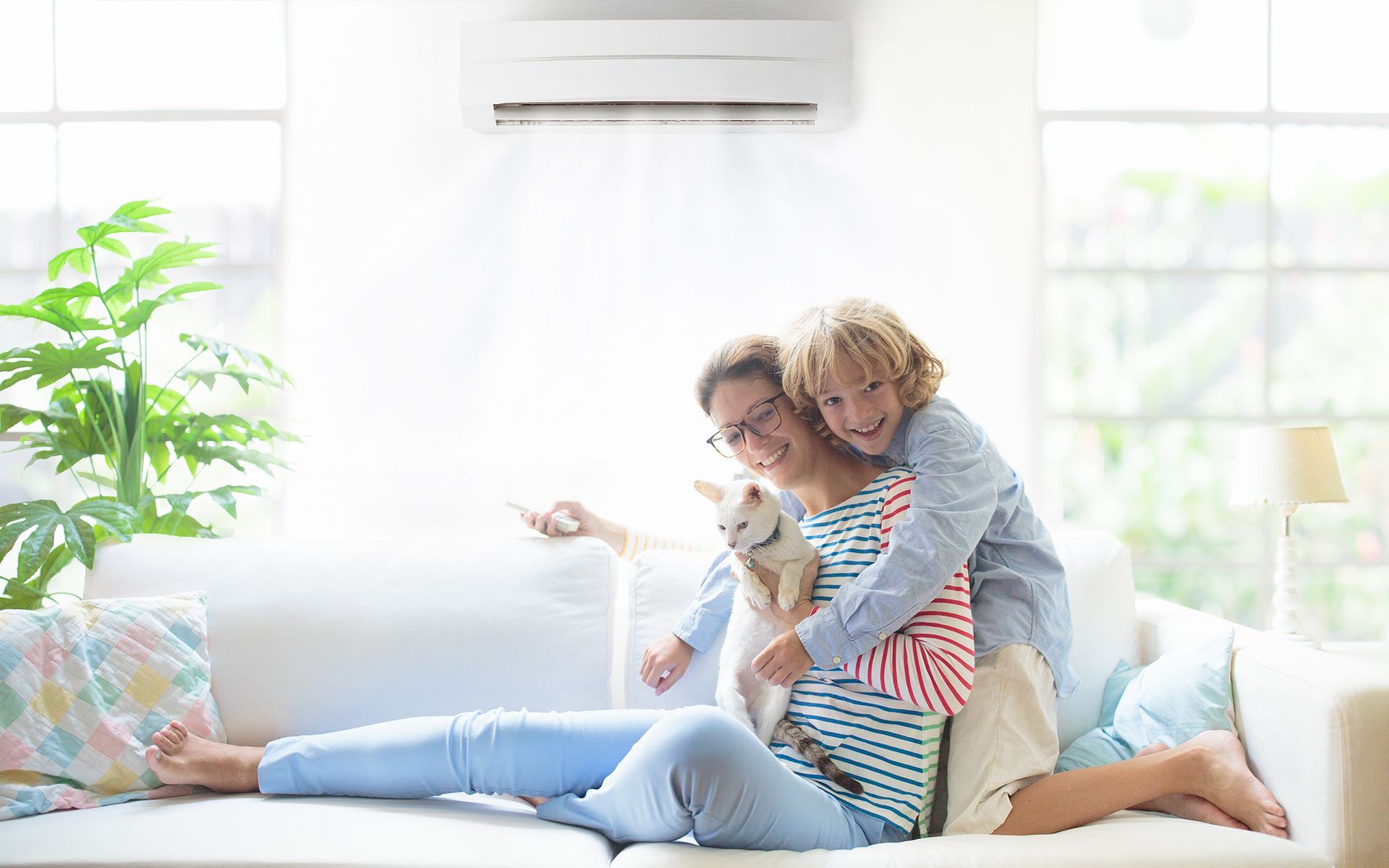 Woman and child on a couch with a cat, under an air conditioner, smiling in a well-lit living room.